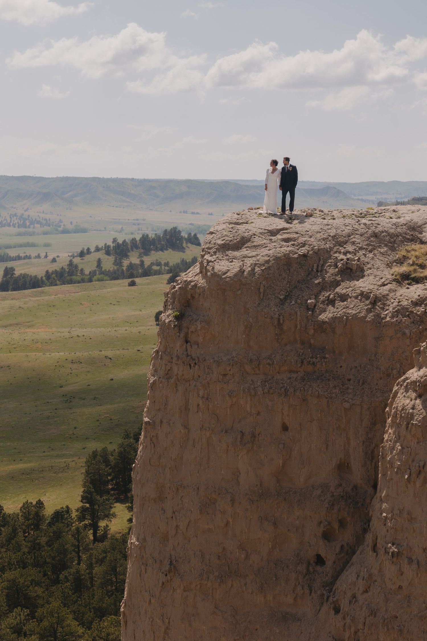 A couple stands on the edge of a dramatic cliff overlooking a vast landscape of rolling hills and forests.