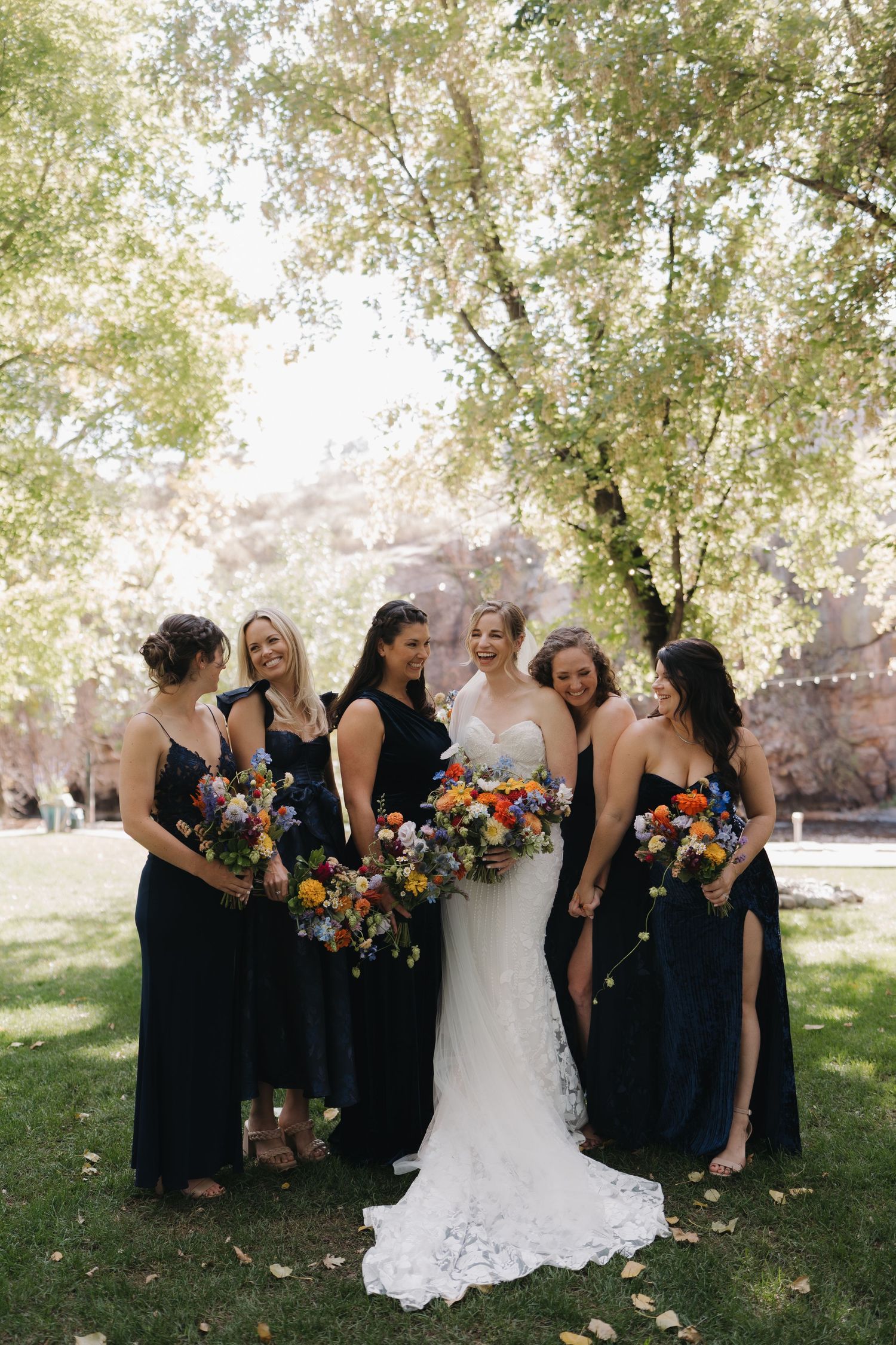 Wedding party in navy dresses and white gown poses under shady trees with colorful bouquets.