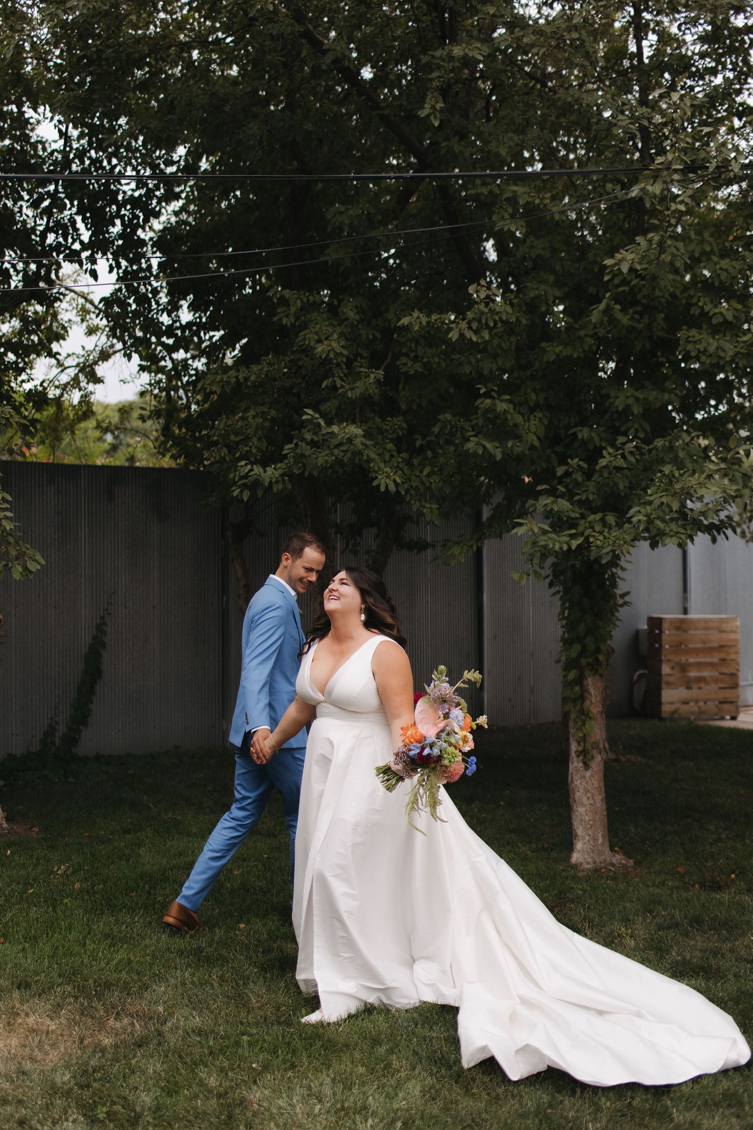 Romantic wedding couple embracing on grassy lawn with flowing white dress and trees in background.