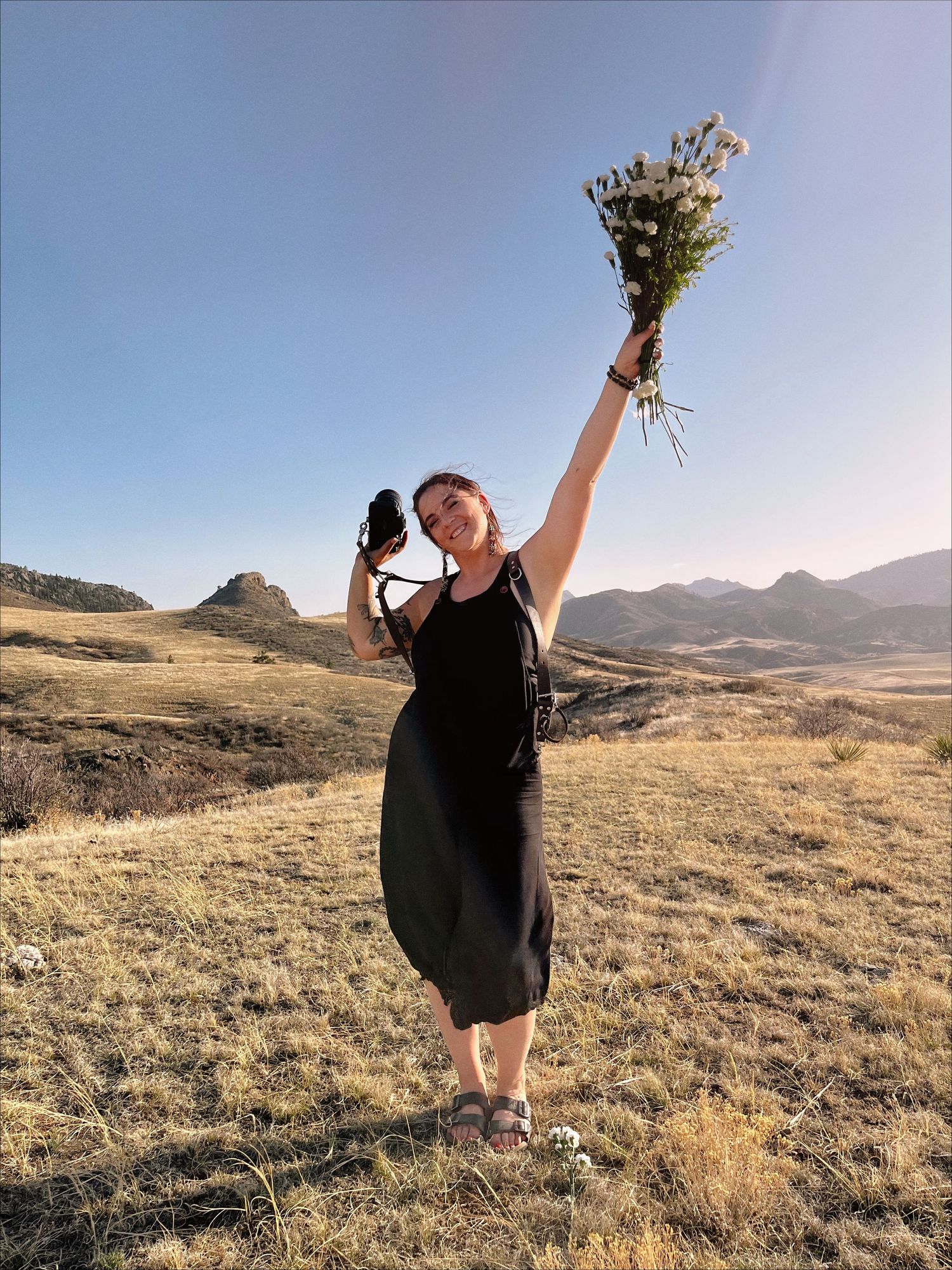 A joyful figure in a black dress holds up a bouquet of dried flowers against a mountainous landscape at sunset.