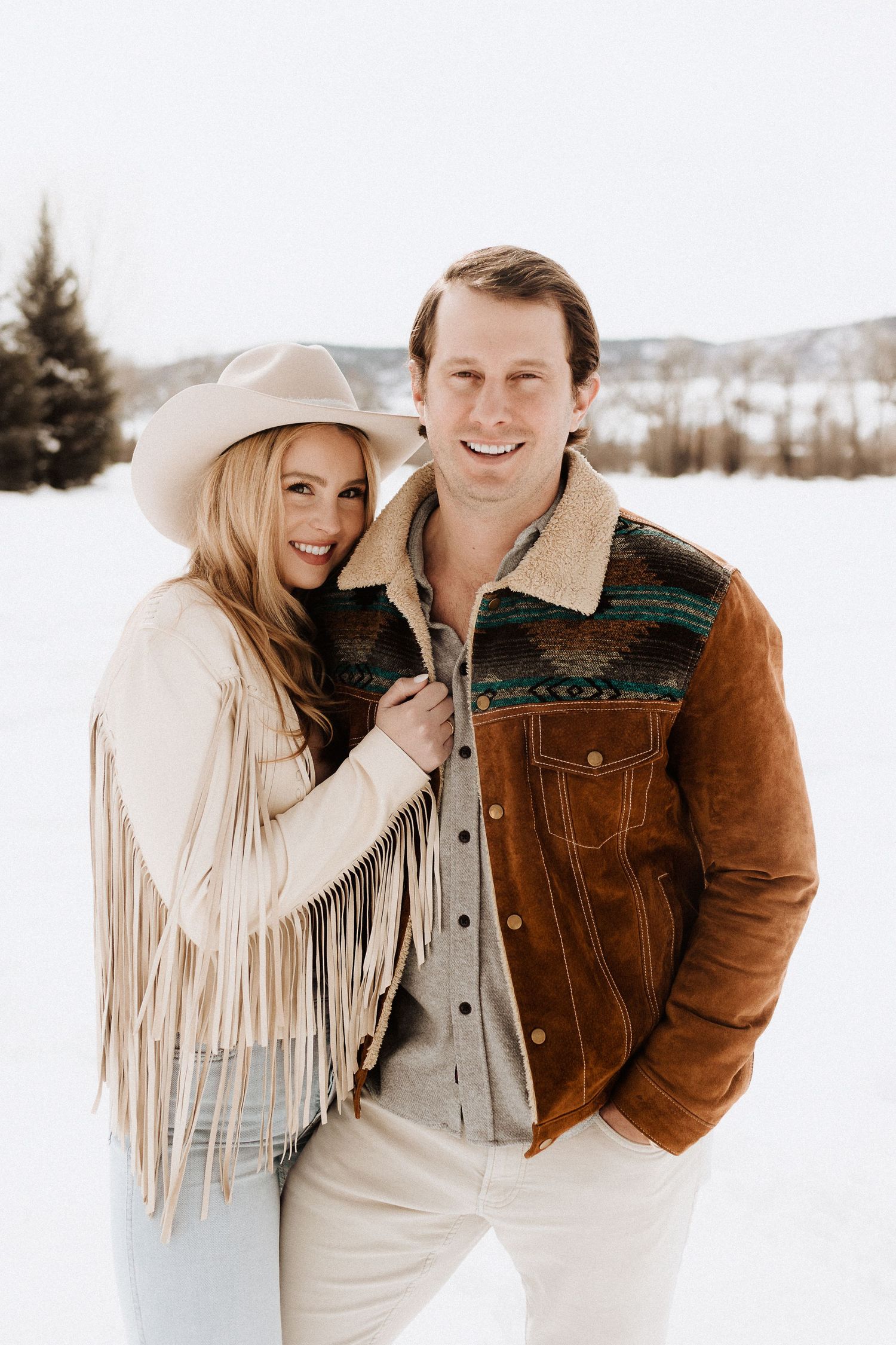 Couple in winter attire with fringe jacket laughing together in snow.