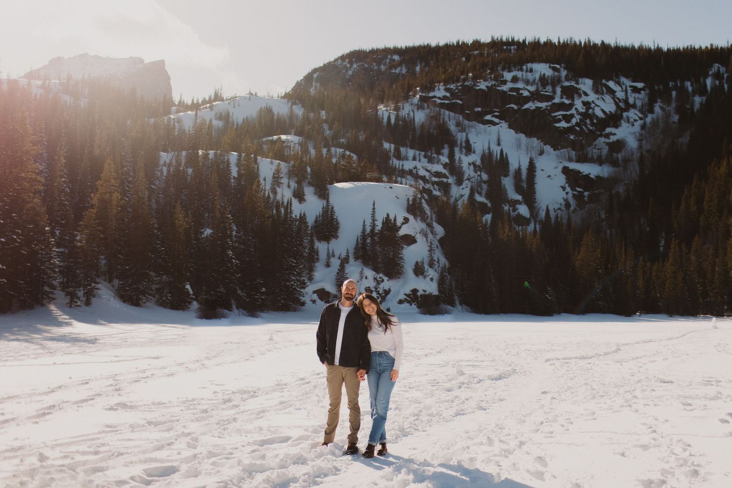 Couple walking together on snowy mountain slope with evergreen forest backdrop.