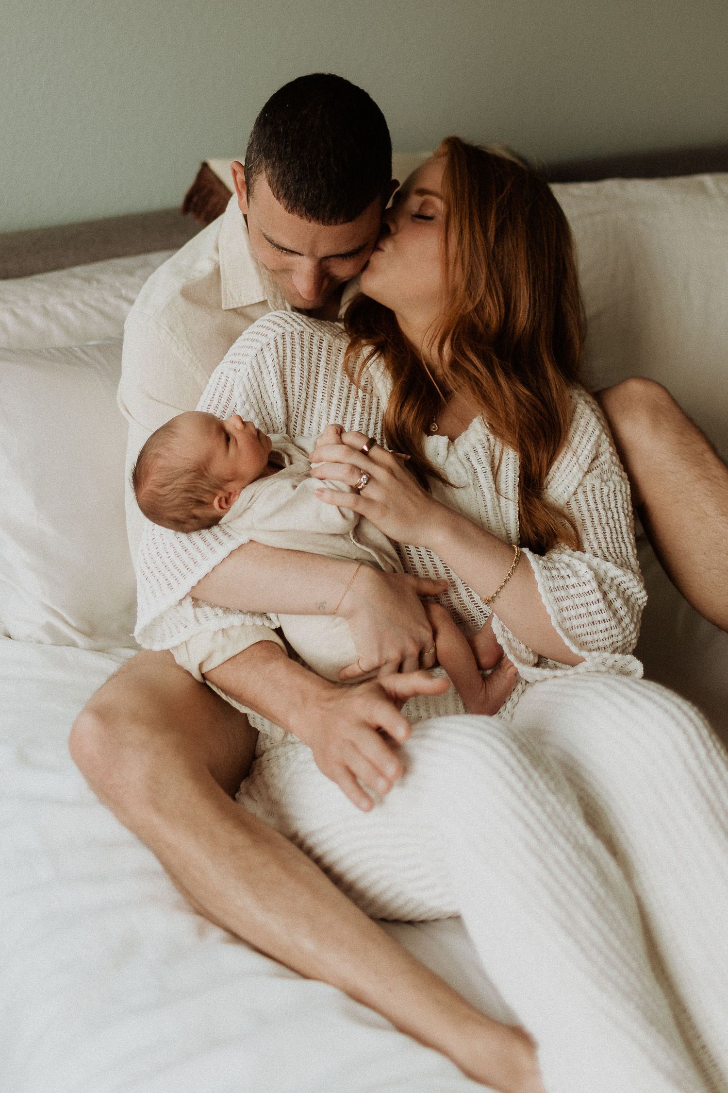 A tender family moment with a newborn baby on a white bed in soft lighting.