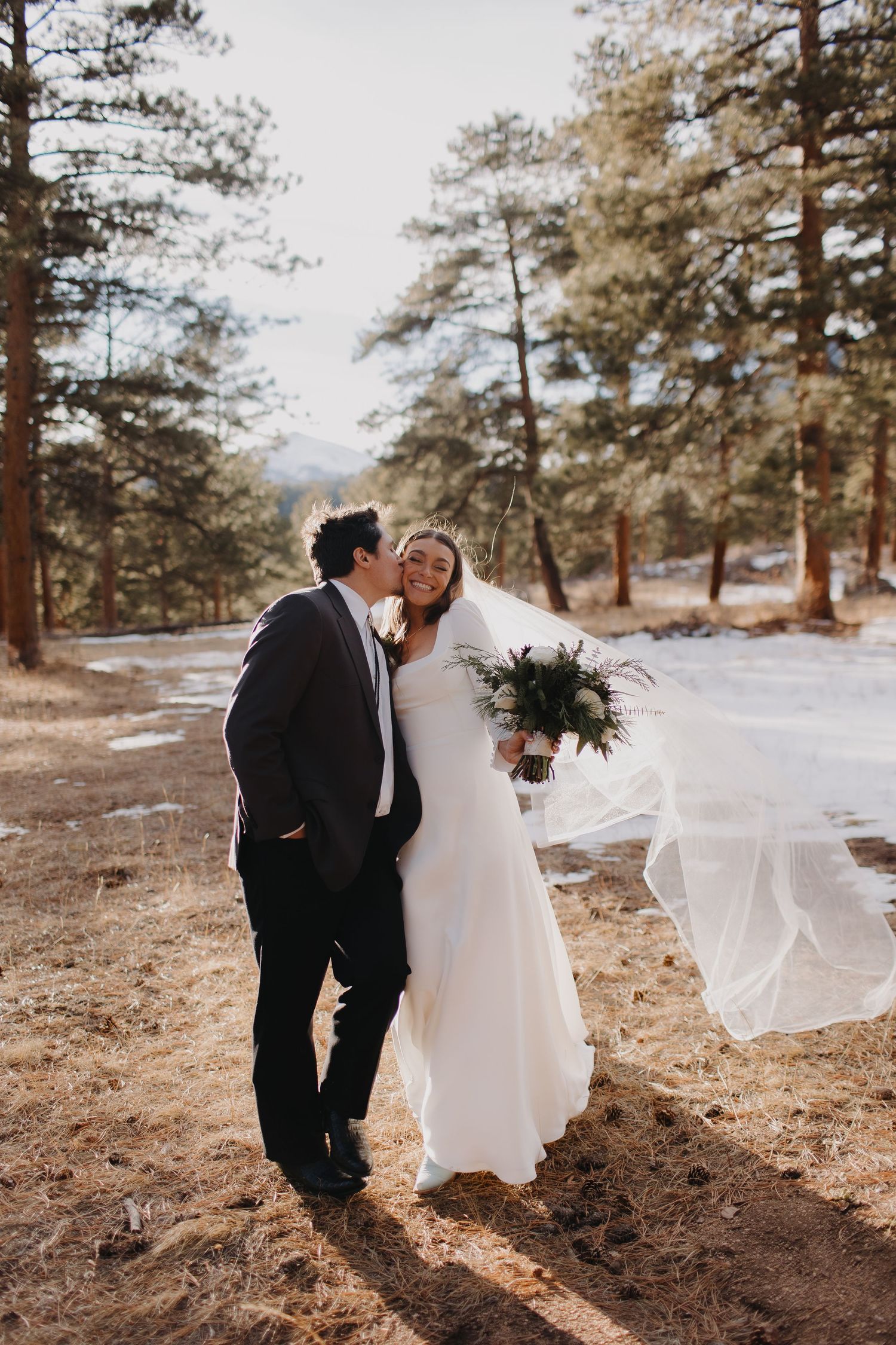 A romantic winter wedding portrait with pine trees and flowing veil in golden sunlight.