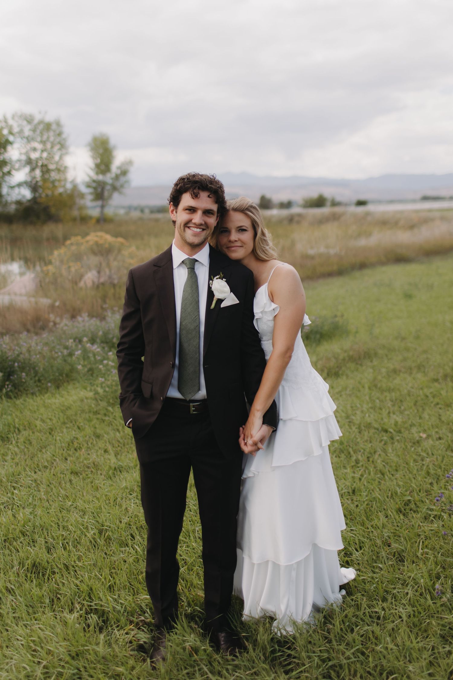 A wedding couple poses together in formal attire on grassy terrain with mountains in the distance.