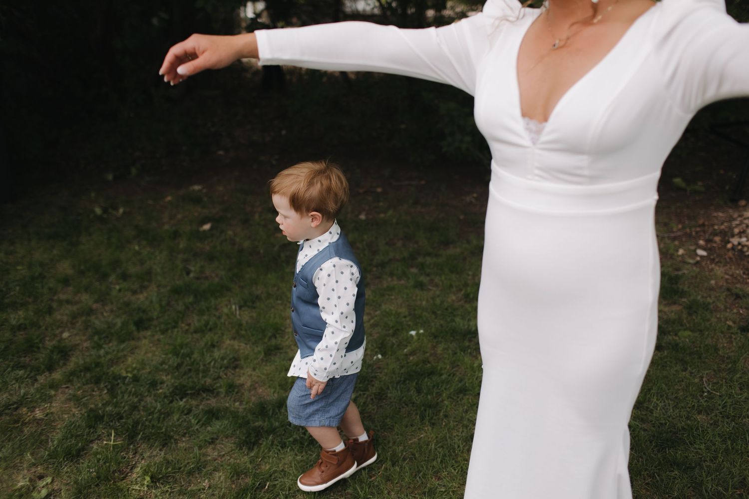 A child in overalls plays outside next to someone in an elegant white wedding dress.