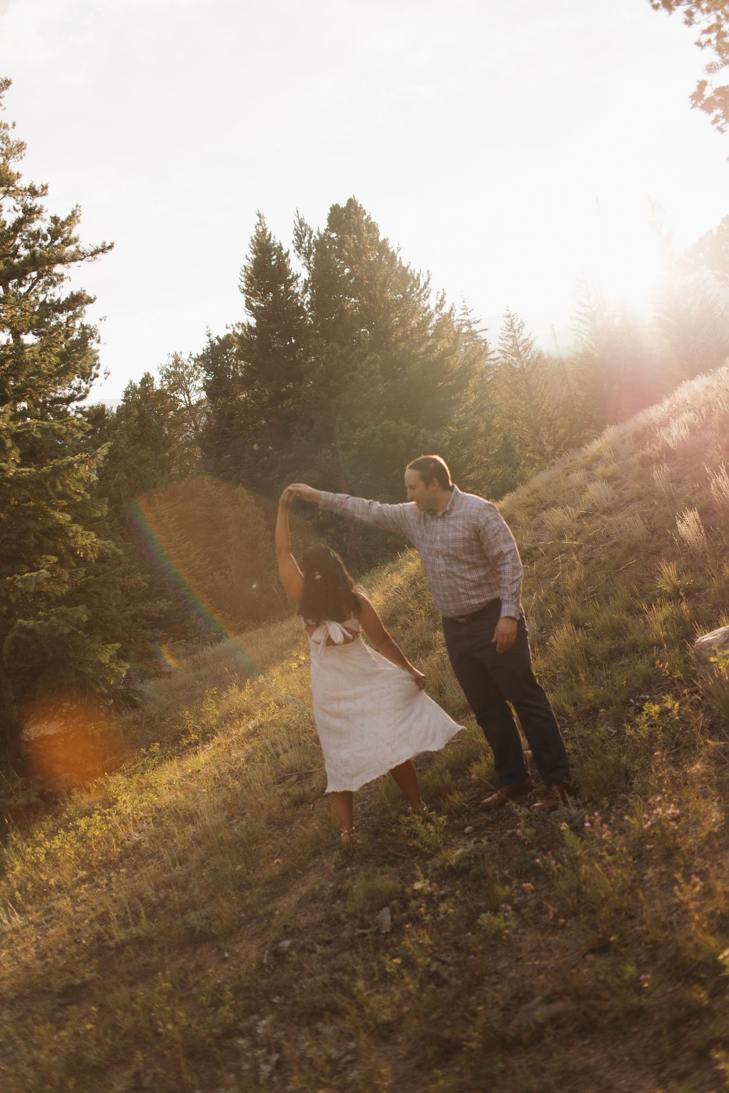 Couple dancing and twirling on a sunlit hillside among pine trees at golden hour.
