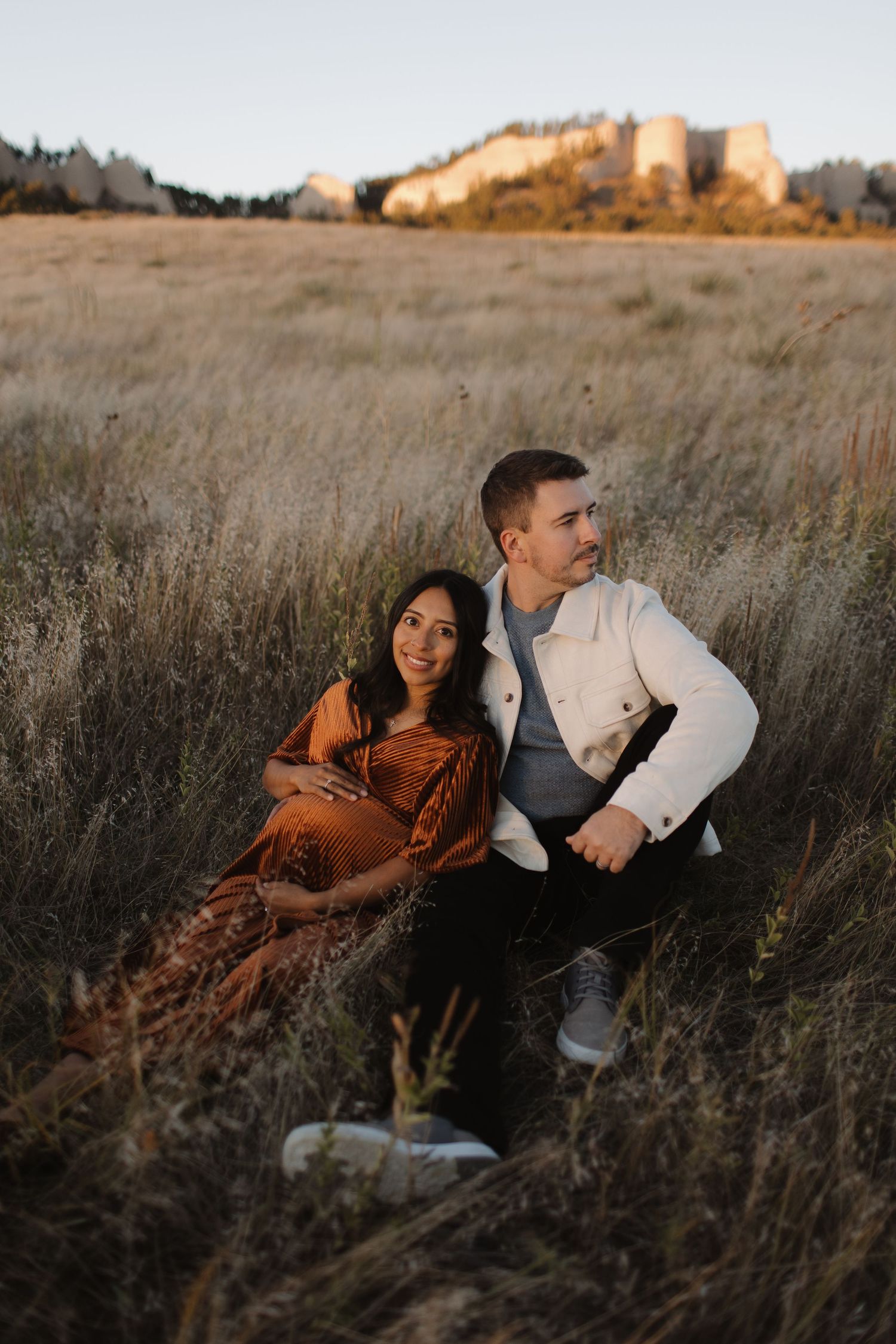 Couple sitting together in tall grass at sunset with rocky cliffs in background.