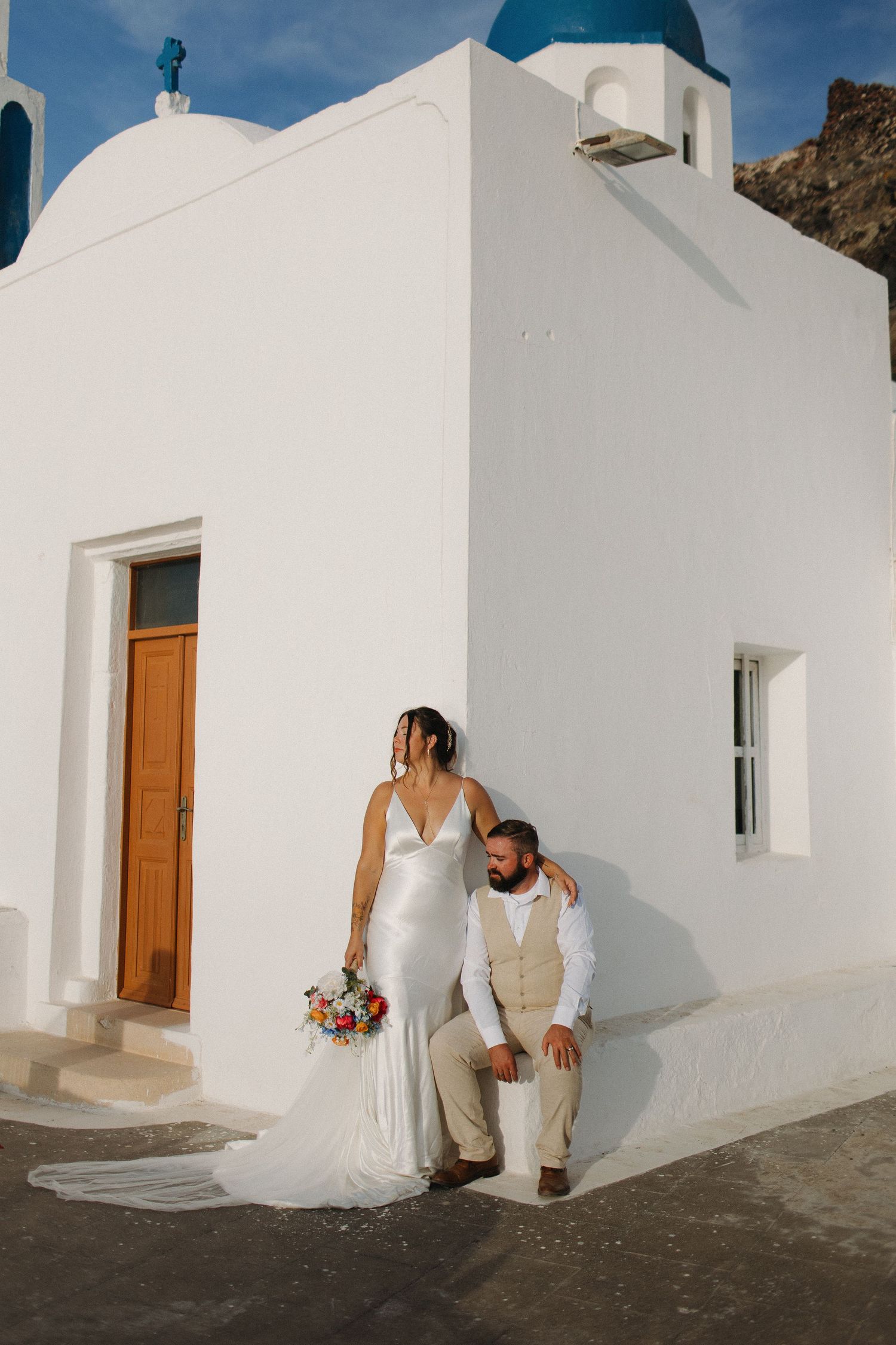 Wedding couple poses against white Mediterranean architecture with blue domed church.
