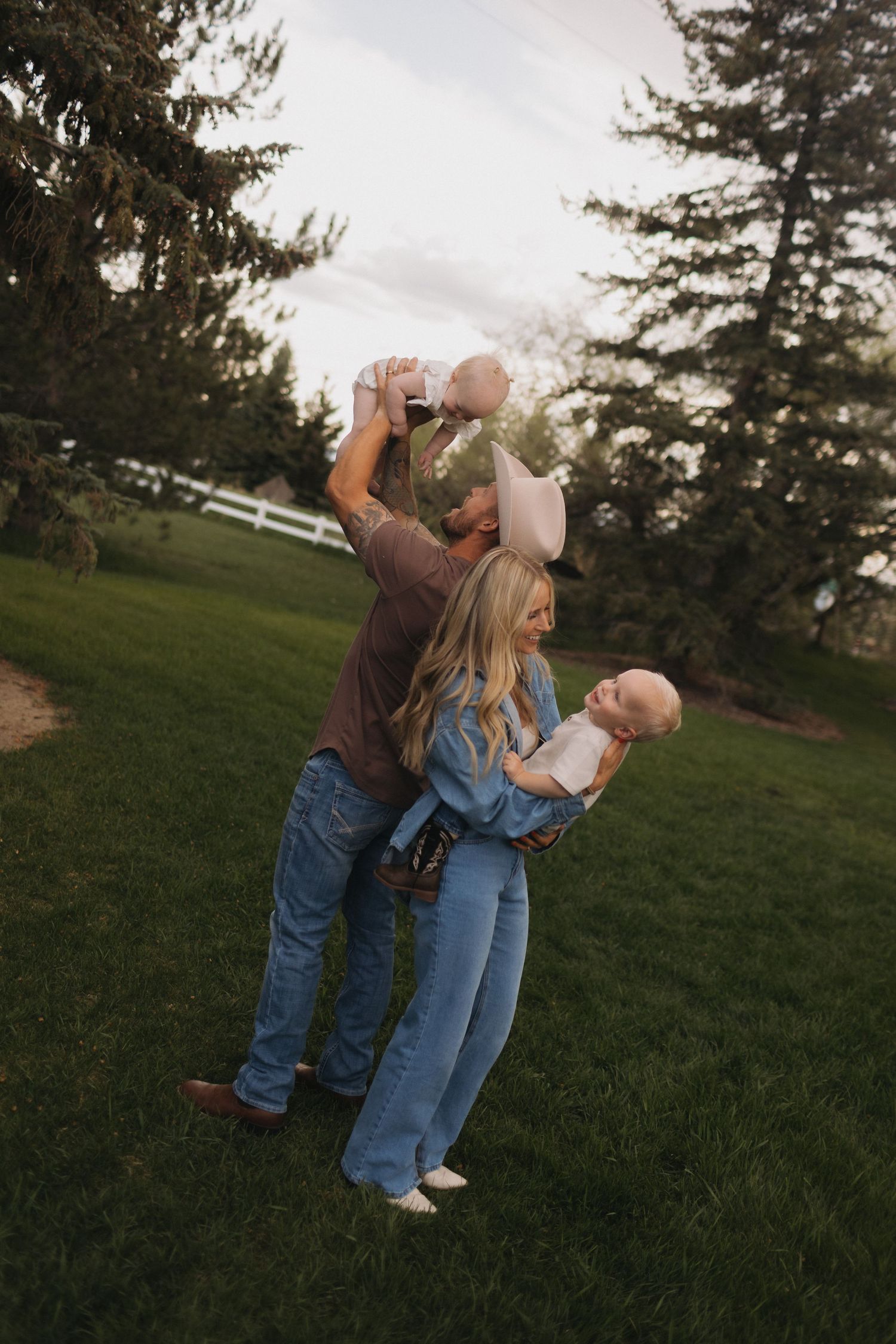Playful family moment lifting babies in air against evergreen trees at sunset.