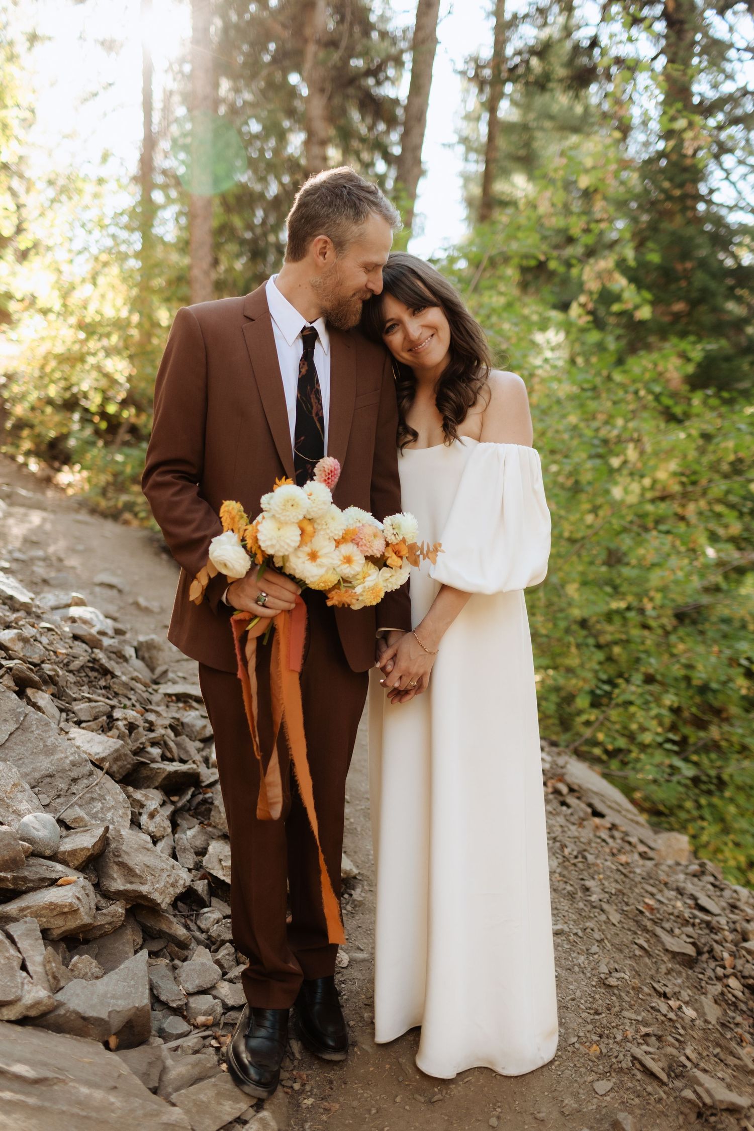 Couple in brown suit and white dress share romantic moments among pine trees.