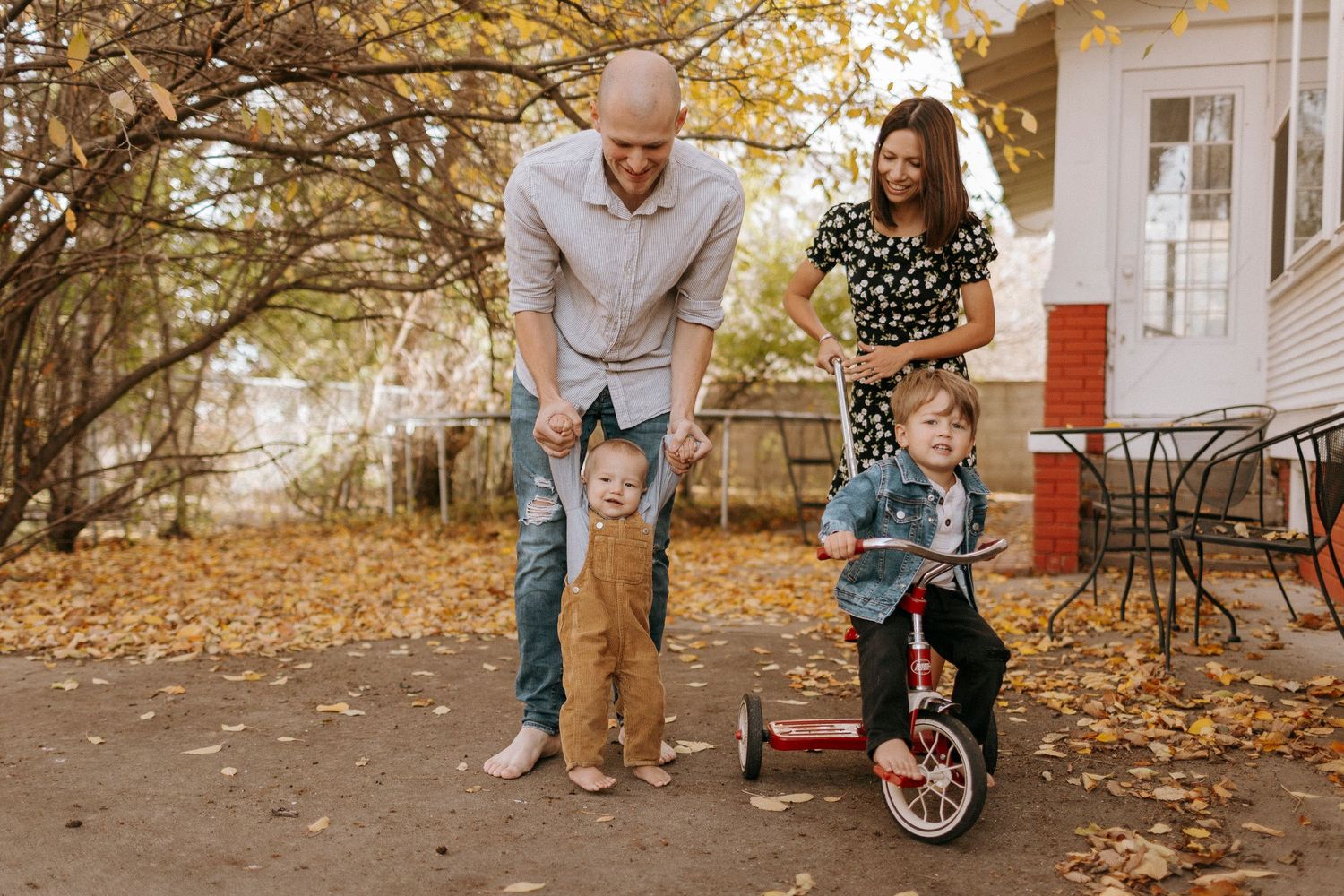 Playful autumn family photoshoot with a red tricycle and fallen leaves in a backyard setting.