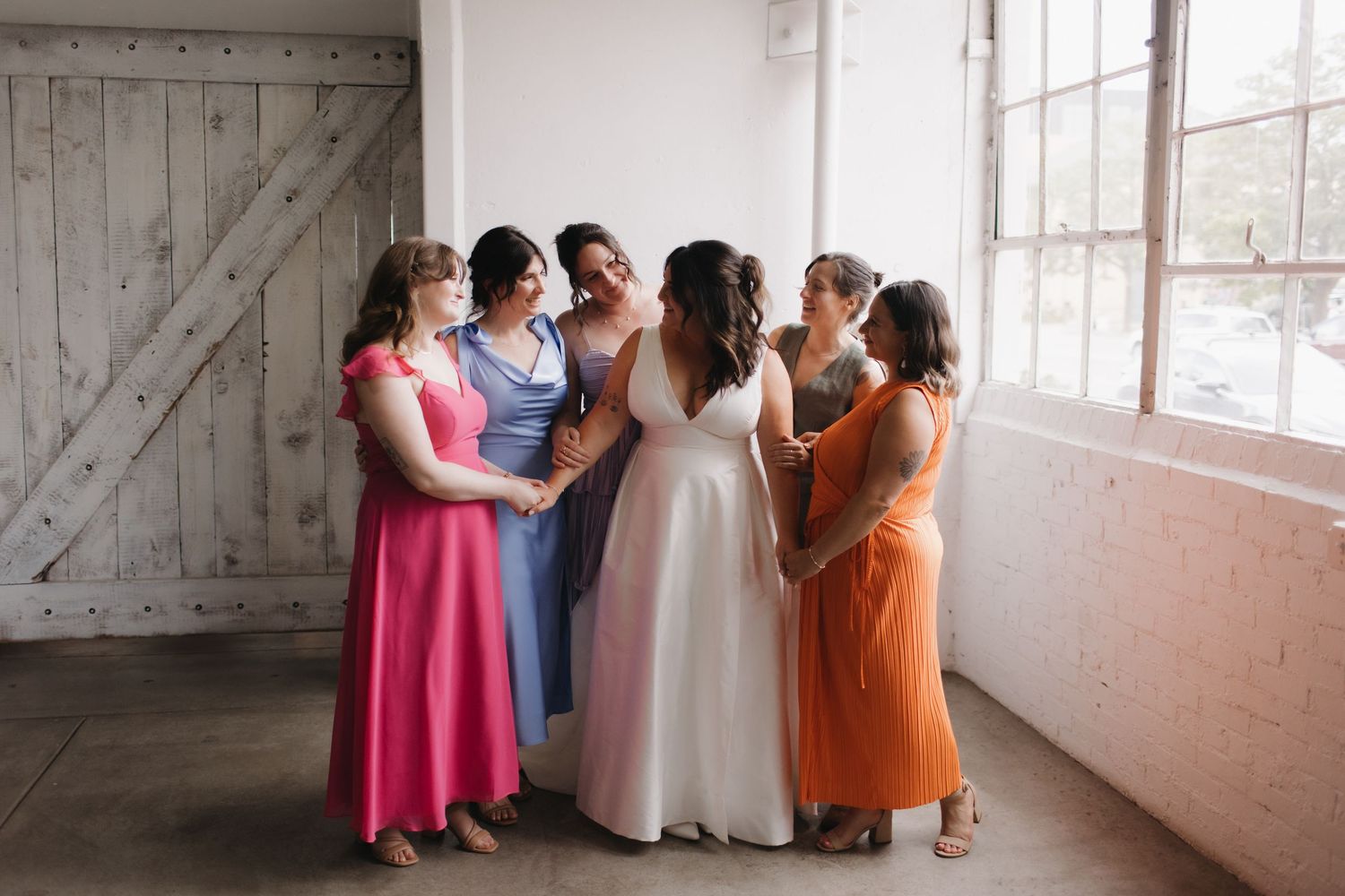 Wedding party members in colorful dresses stand in line against a rustic white wall.