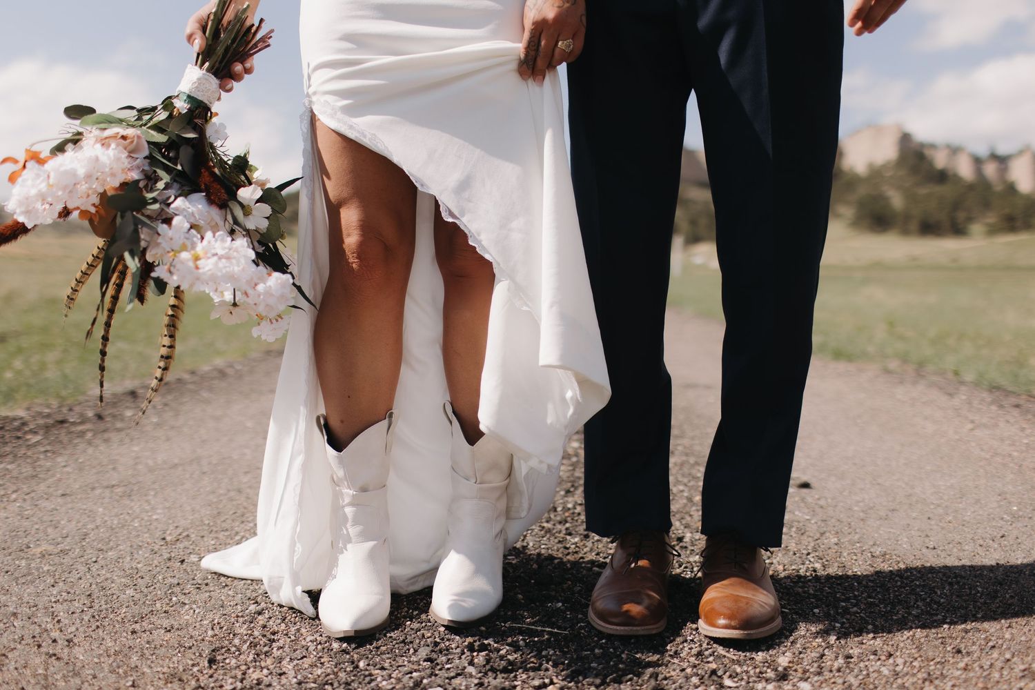 Close-up of wedding attire showing white boots and dress hem next to formal shoes on a path.