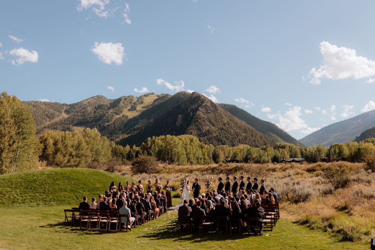 Panoramic mountain landscape with wedding guests seated on wooden benches against scenic backdrop.