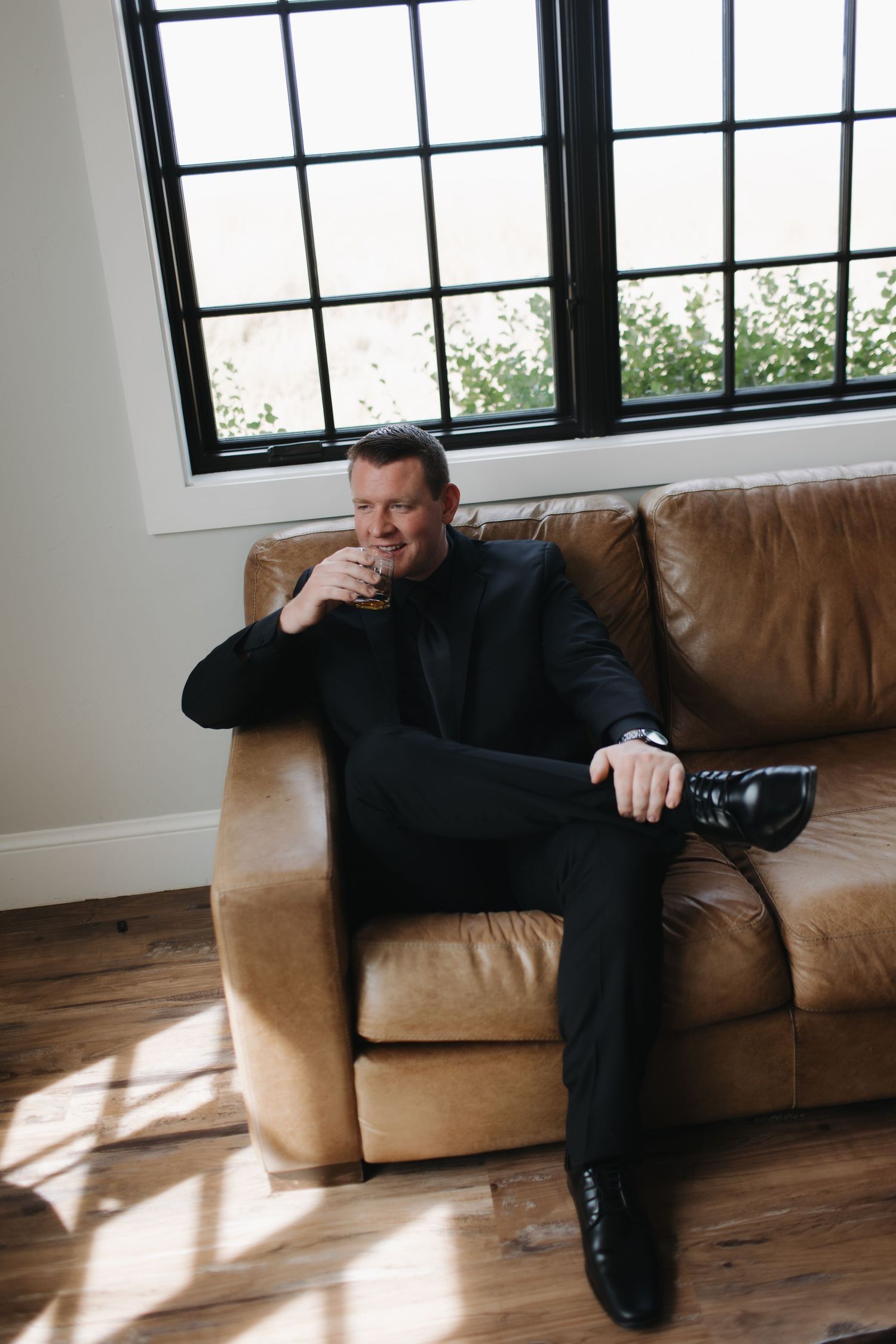 Person in dark suit relaxing on brown leather couch in room with large windows.