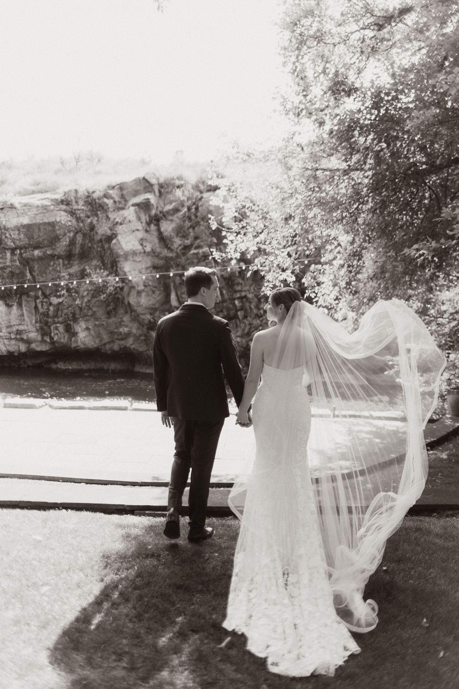 Black and white portrait of wedding couple walking with flowing veil in garden setting.