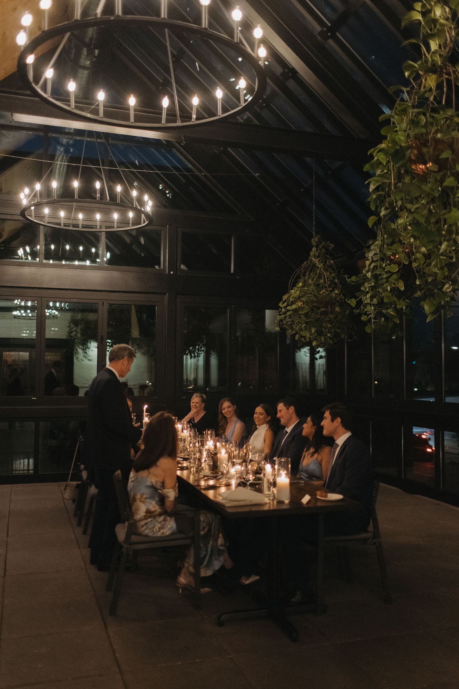 Wedding reception dinner under string lights in modern glass conservatory at night.