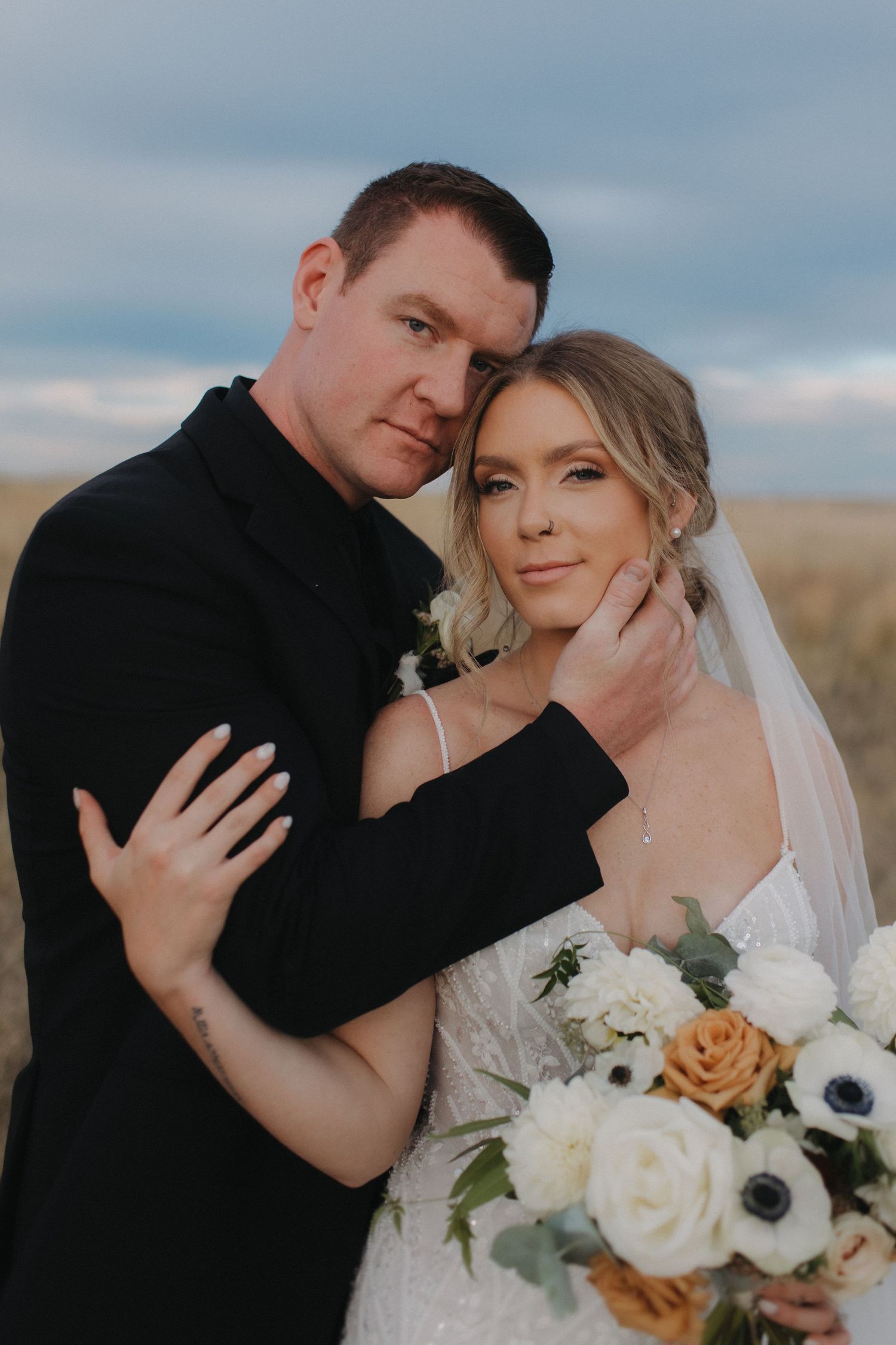 Intimate wedding portrait with white and peach floral bouquet against cloudy sky.
