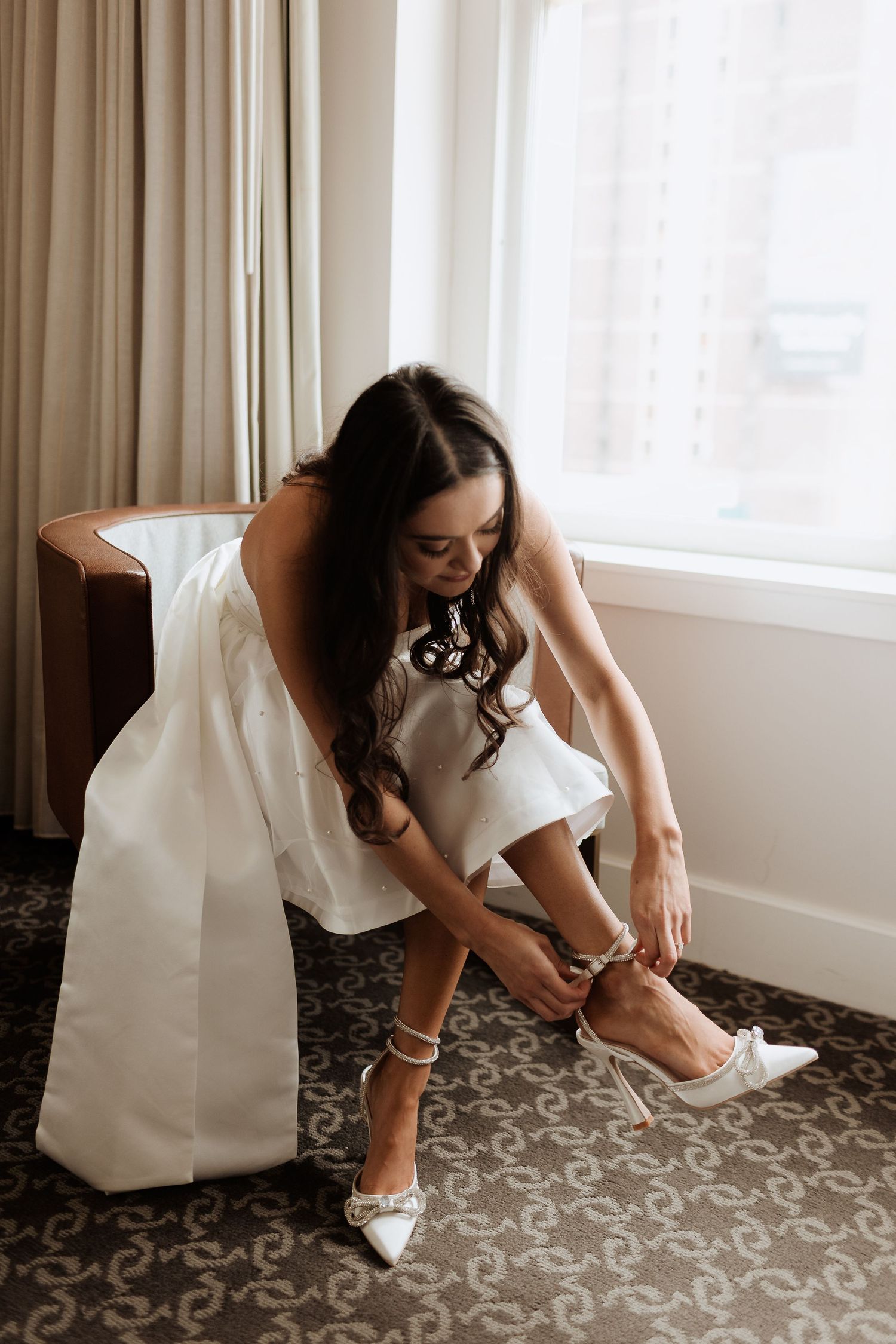 Person in white wedding dress putting on shoes on patterned hotel carpet.