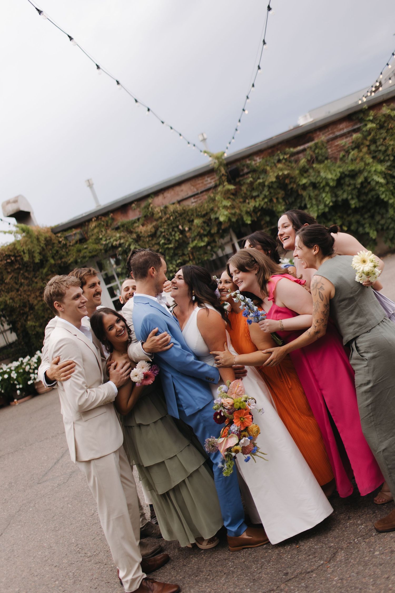 Wedding party lined up outdoors sharing laughs and embraces under string lights.