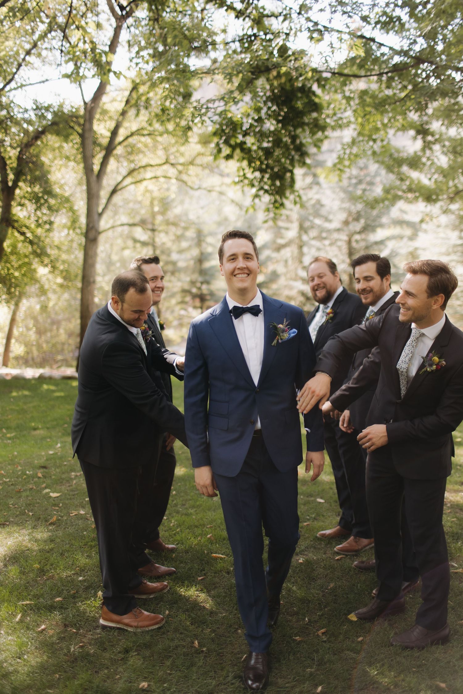 Groomsmen in suits share a candid laughing moment on grass under trees.