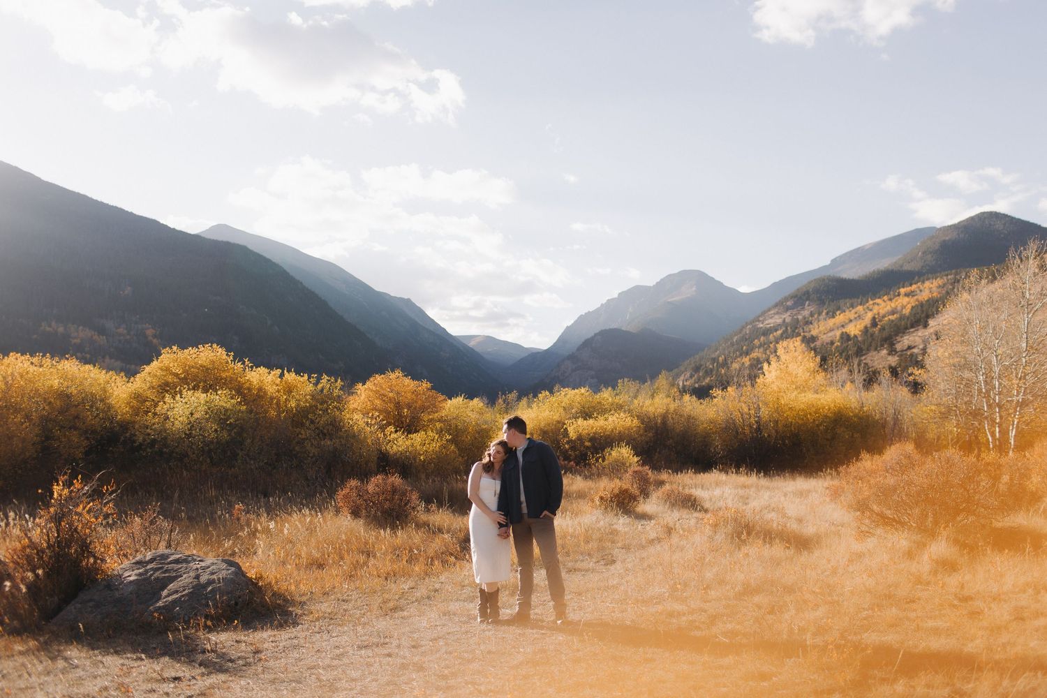 A couple walks through a scenic mountain landscape with golden autumn foliage and dramatic cloudy skies.
