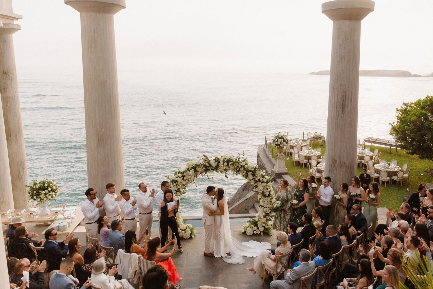 Outdoor wedding ceremony taking place under white pillars overlooking the ocean with guests seated in rows.