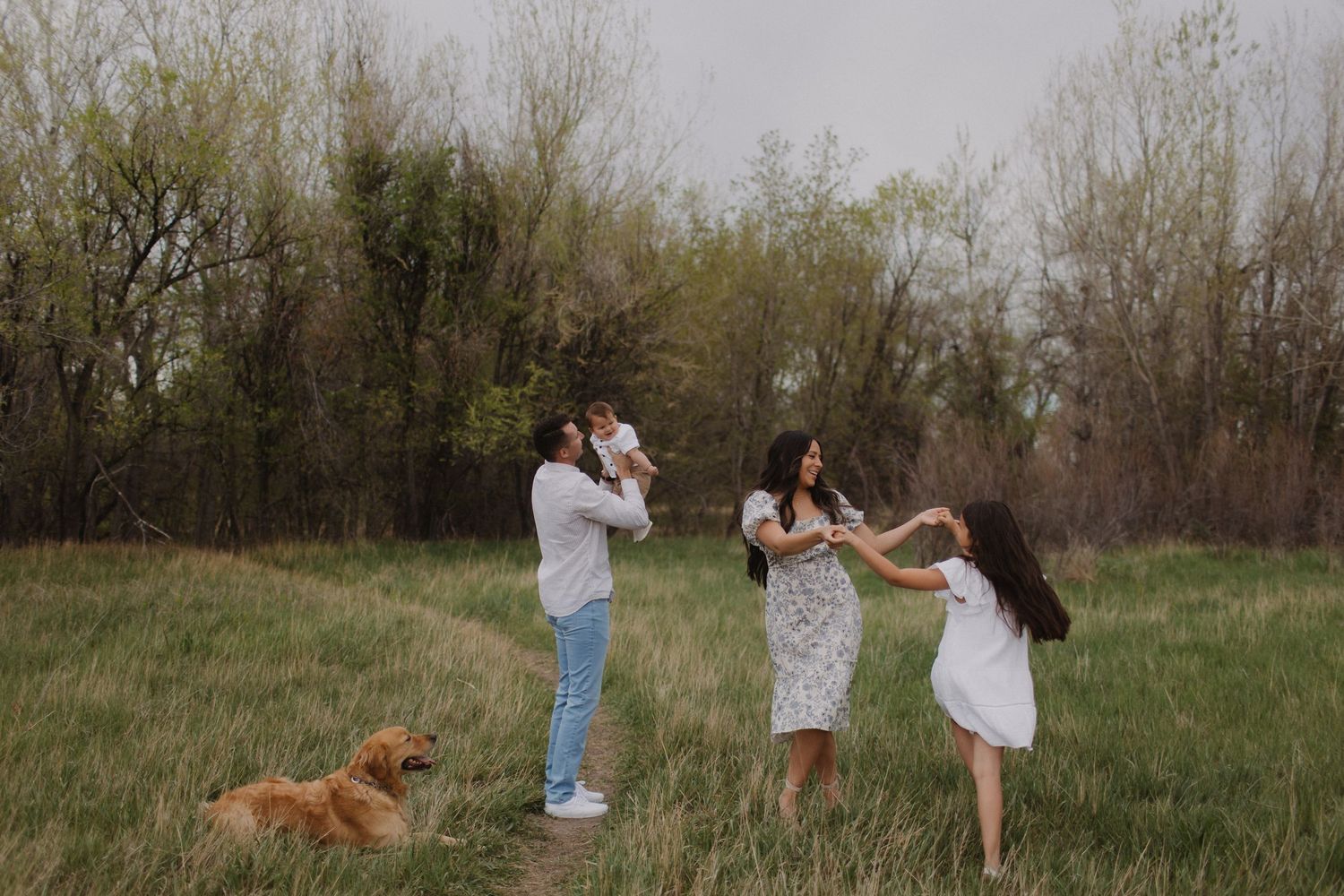 Family playing and dancing together in a grassy field with a dog running alongside.