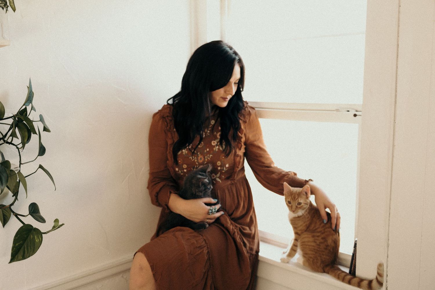 A woman in a brown dress pets a cat in a bright indoor setting with houseplants.