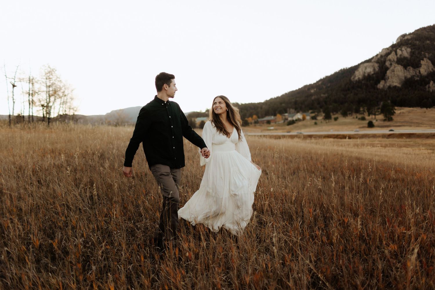 Couple walking through golden prairie grass with mountains and church in background.