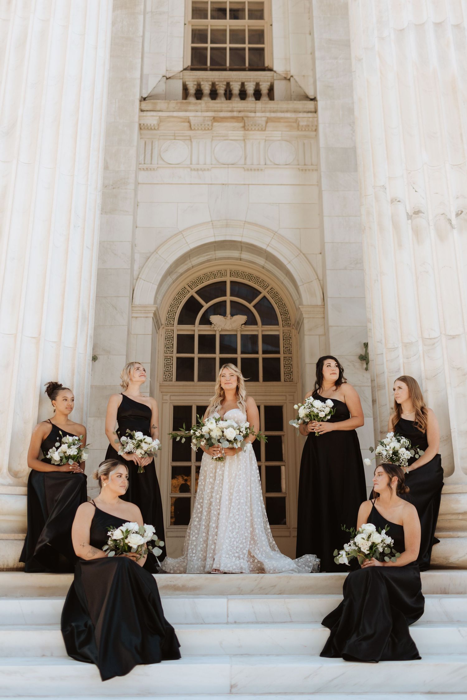 Wedding party in black dresses poses elegantly against white columned building.