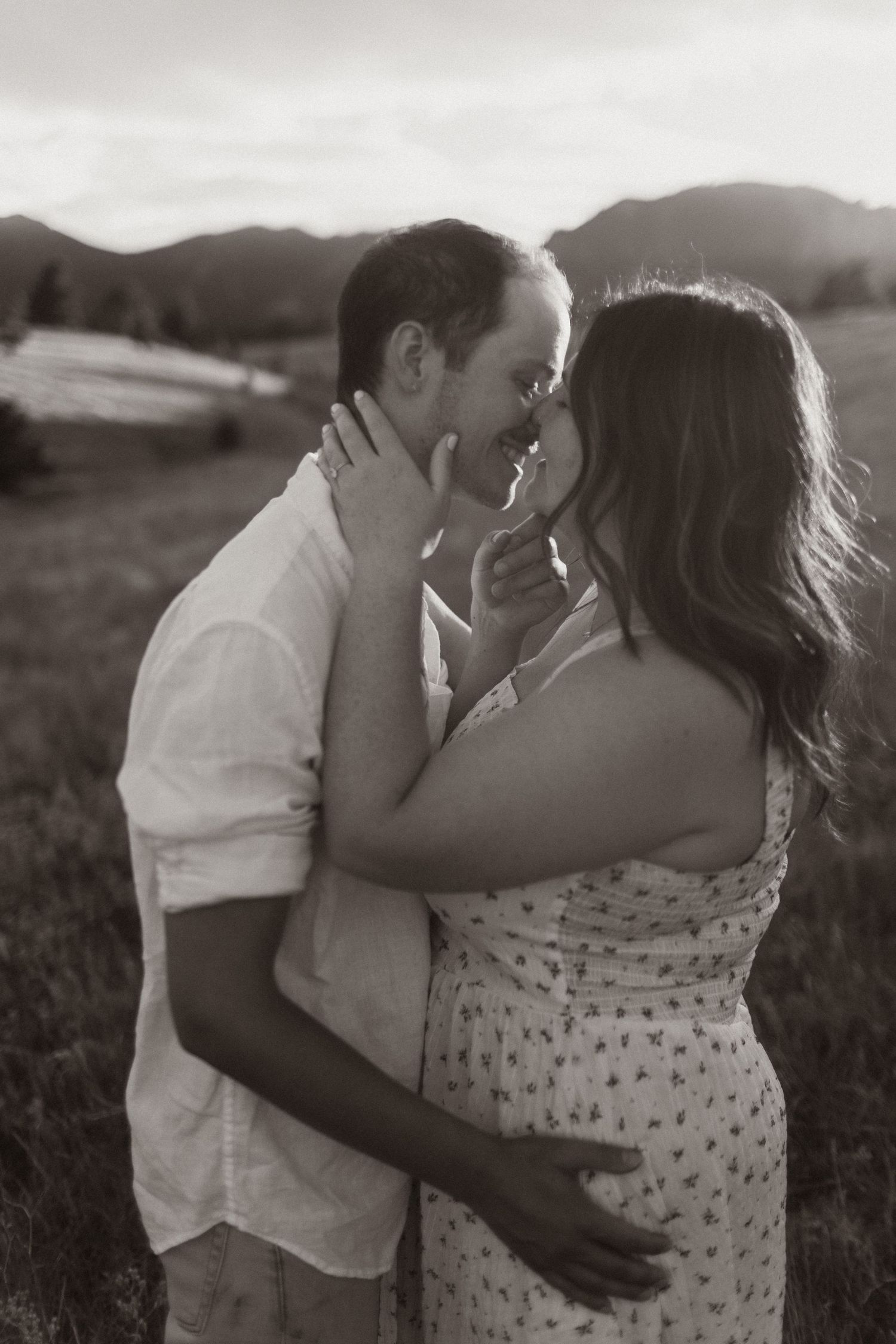 A romantic black and white portrait of a couple embracing in a field at sunset.