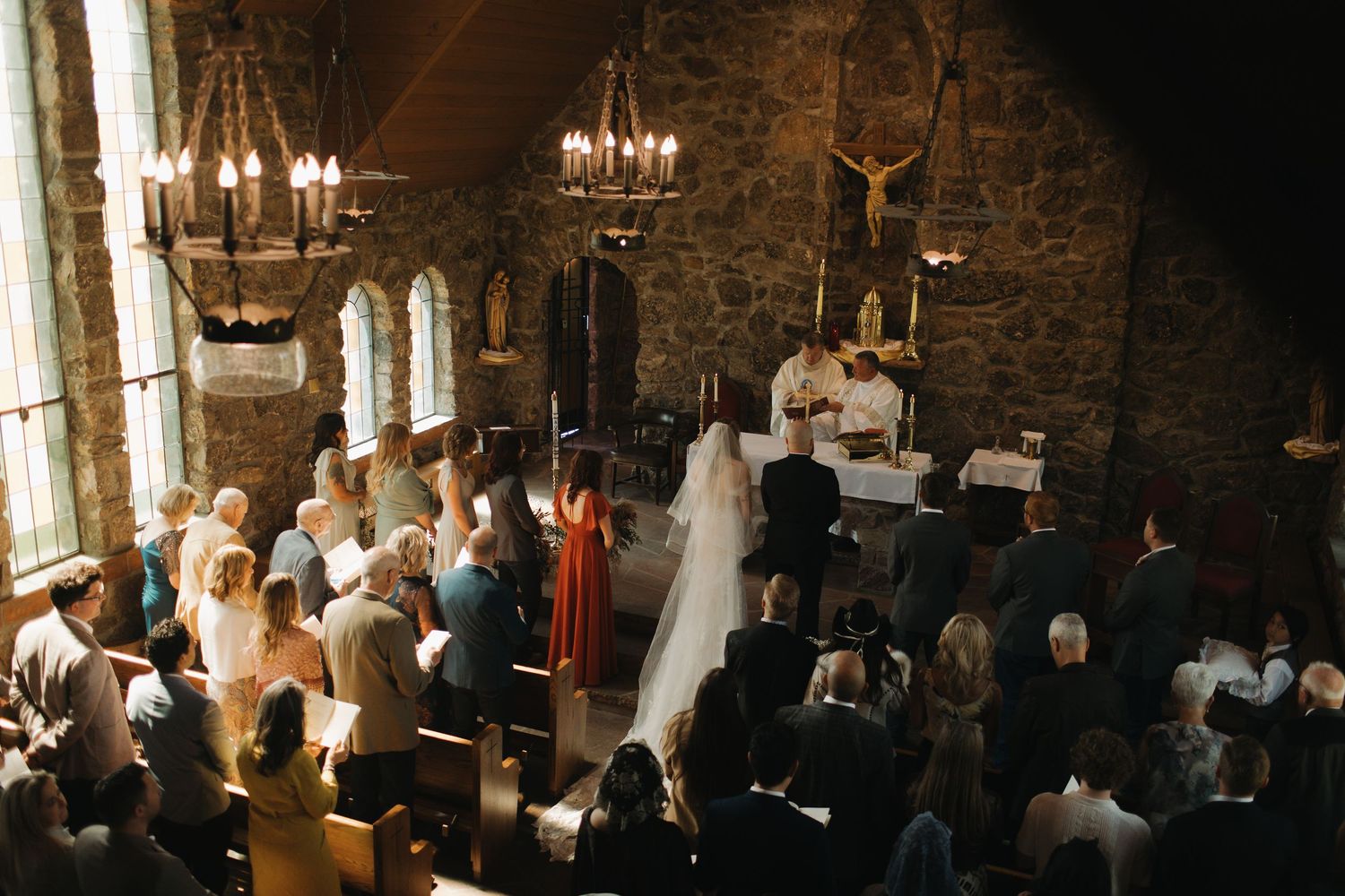 Elegant wedding ceremony in a rustic stone venue with chandeliers and natural light streaming through windows.