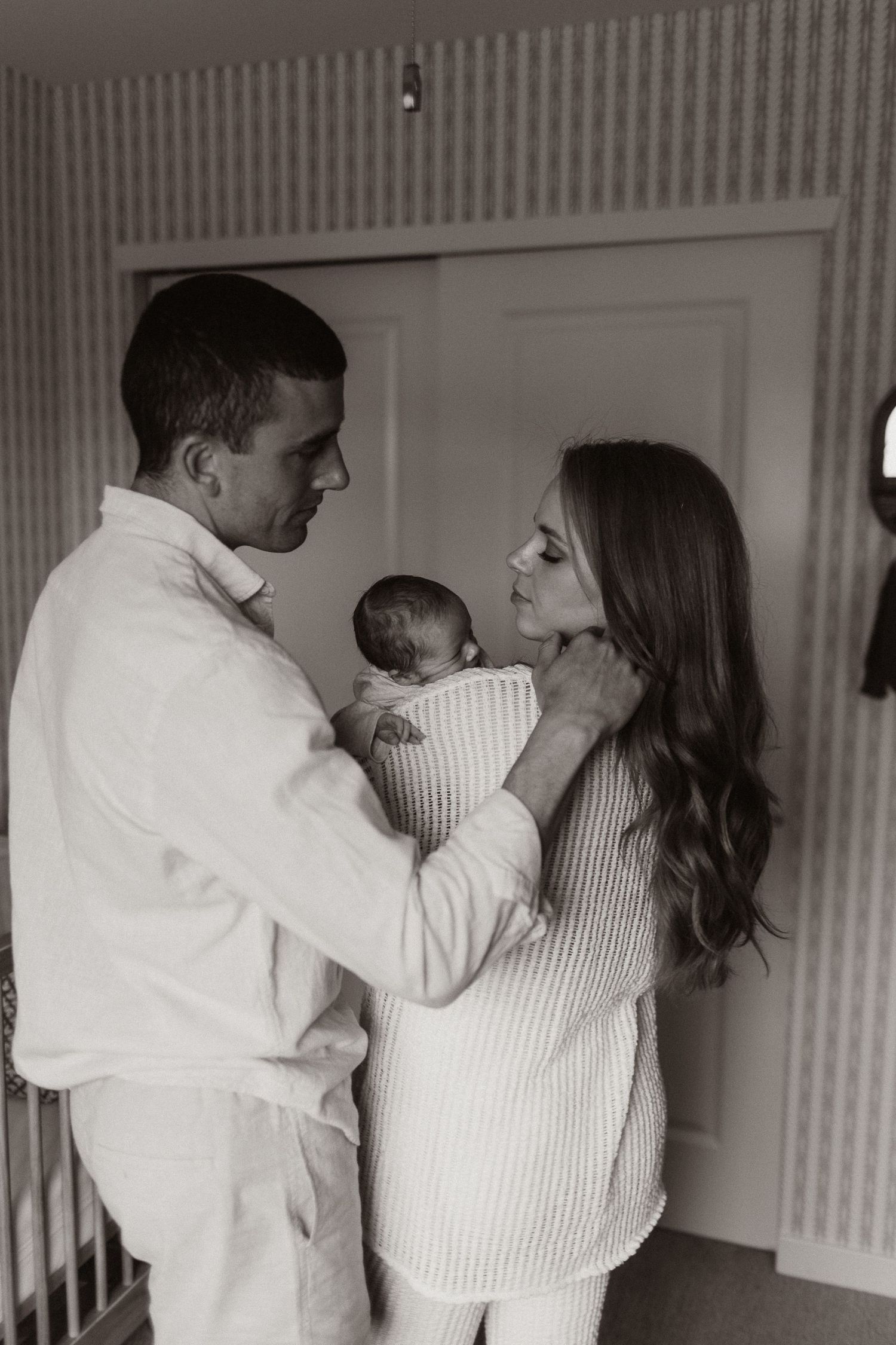 A couple in white clothing shares an intimate moment with their newborn baby against striped wallpaper.