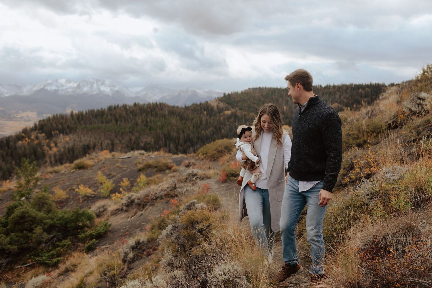 A family hikes together on an autumn mountain trail with snow-capped peaks in the distance.