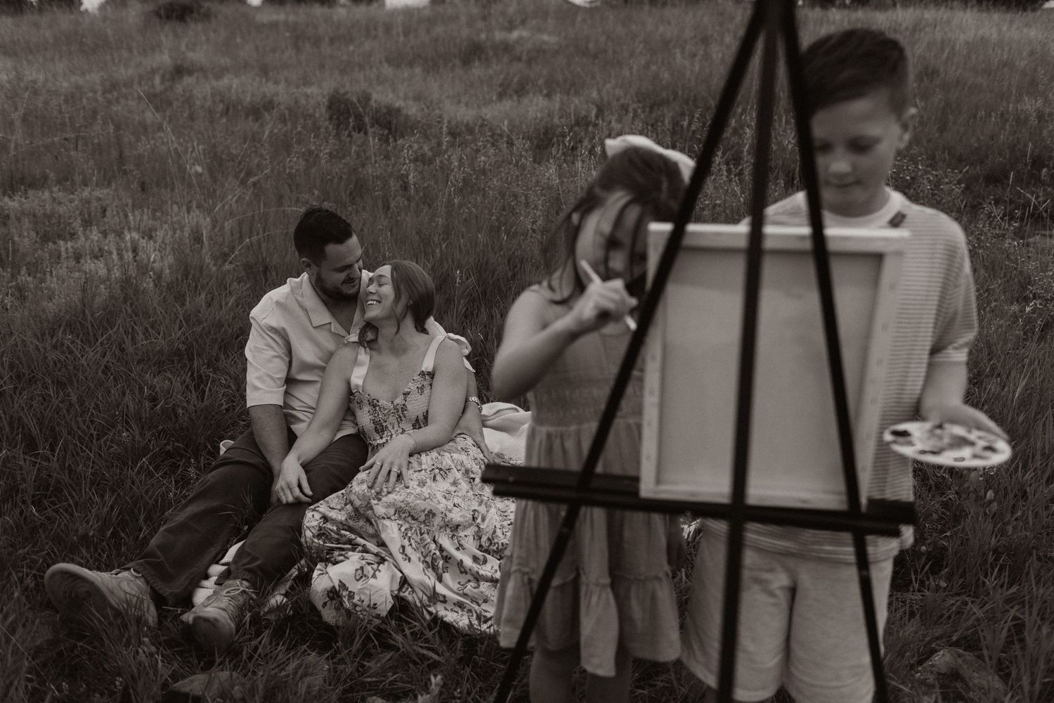 Family gathering around an art easel in a rustic outdoor setting.