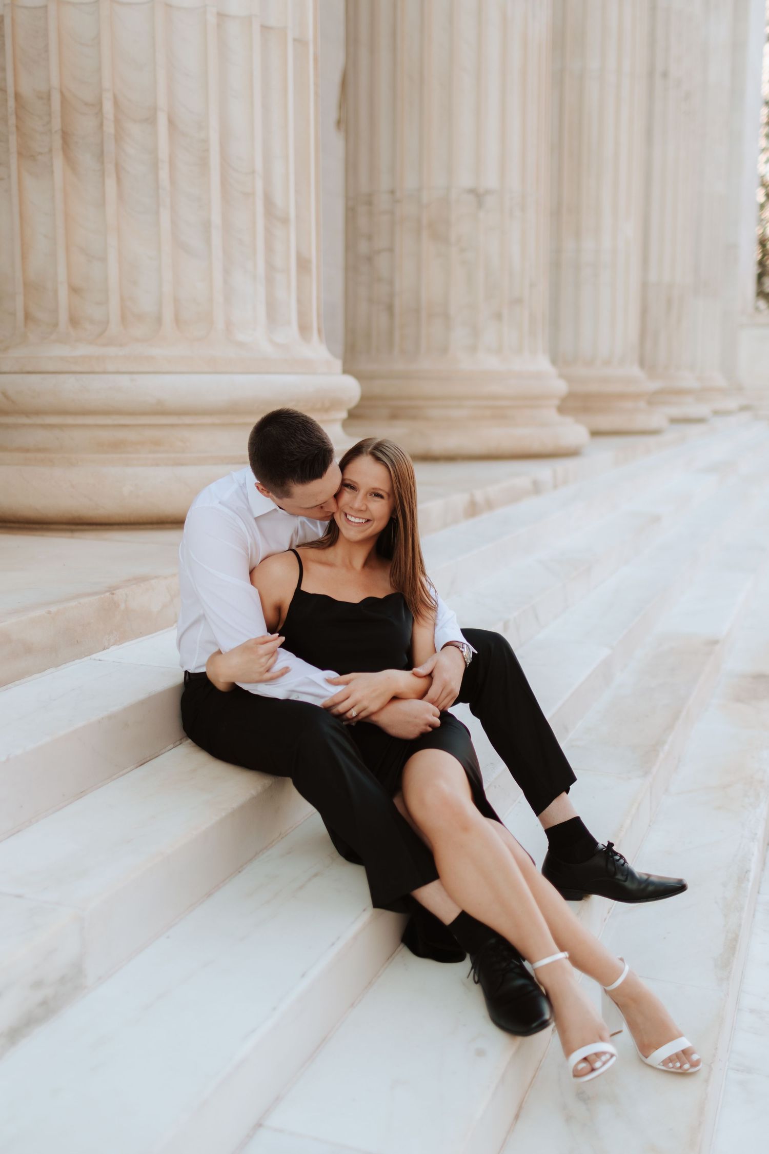 Playful couple lounging on white marble steps between elegant columns.