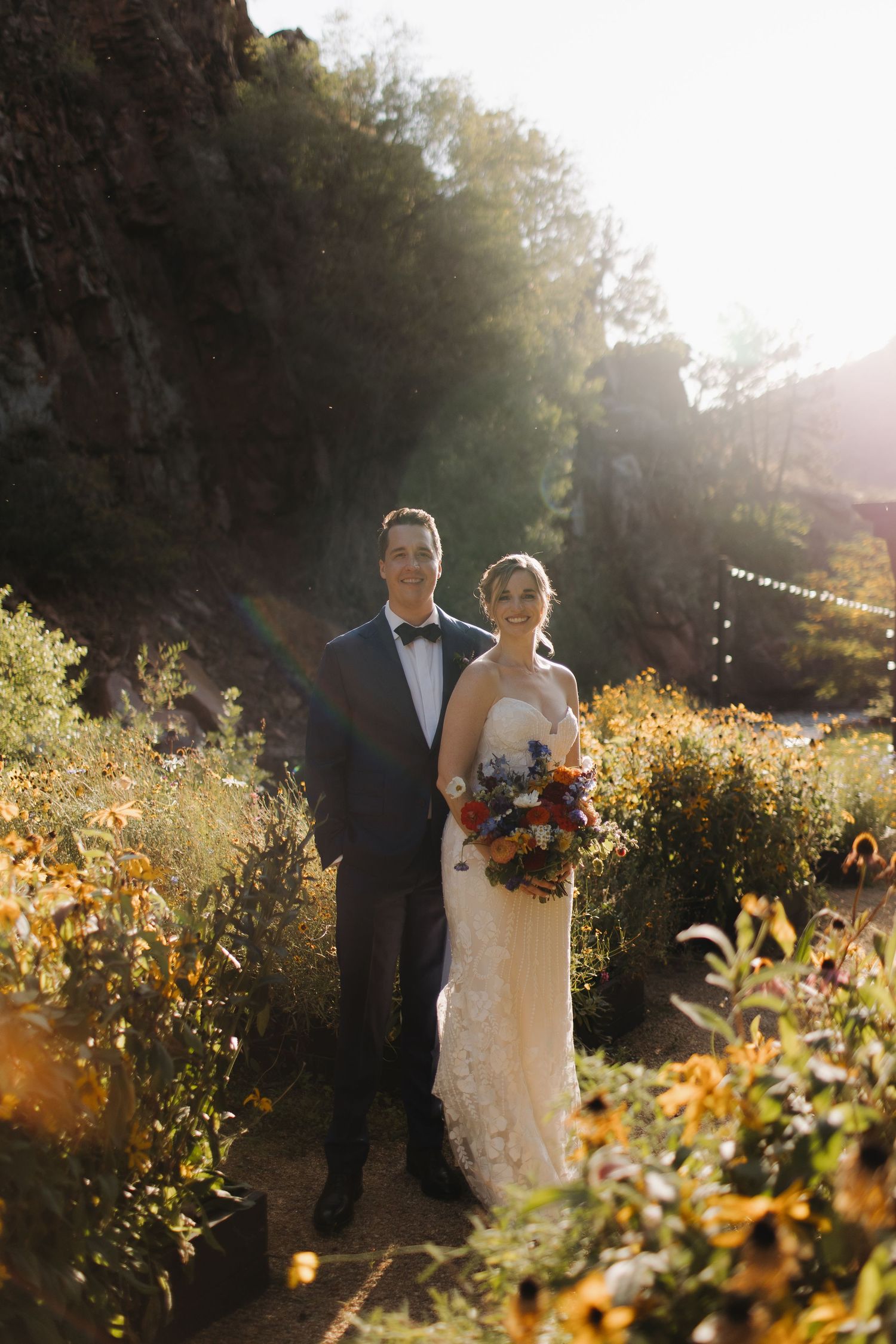 Newlyweds walk through golden sunlit wildflowers during sunset portrait session.