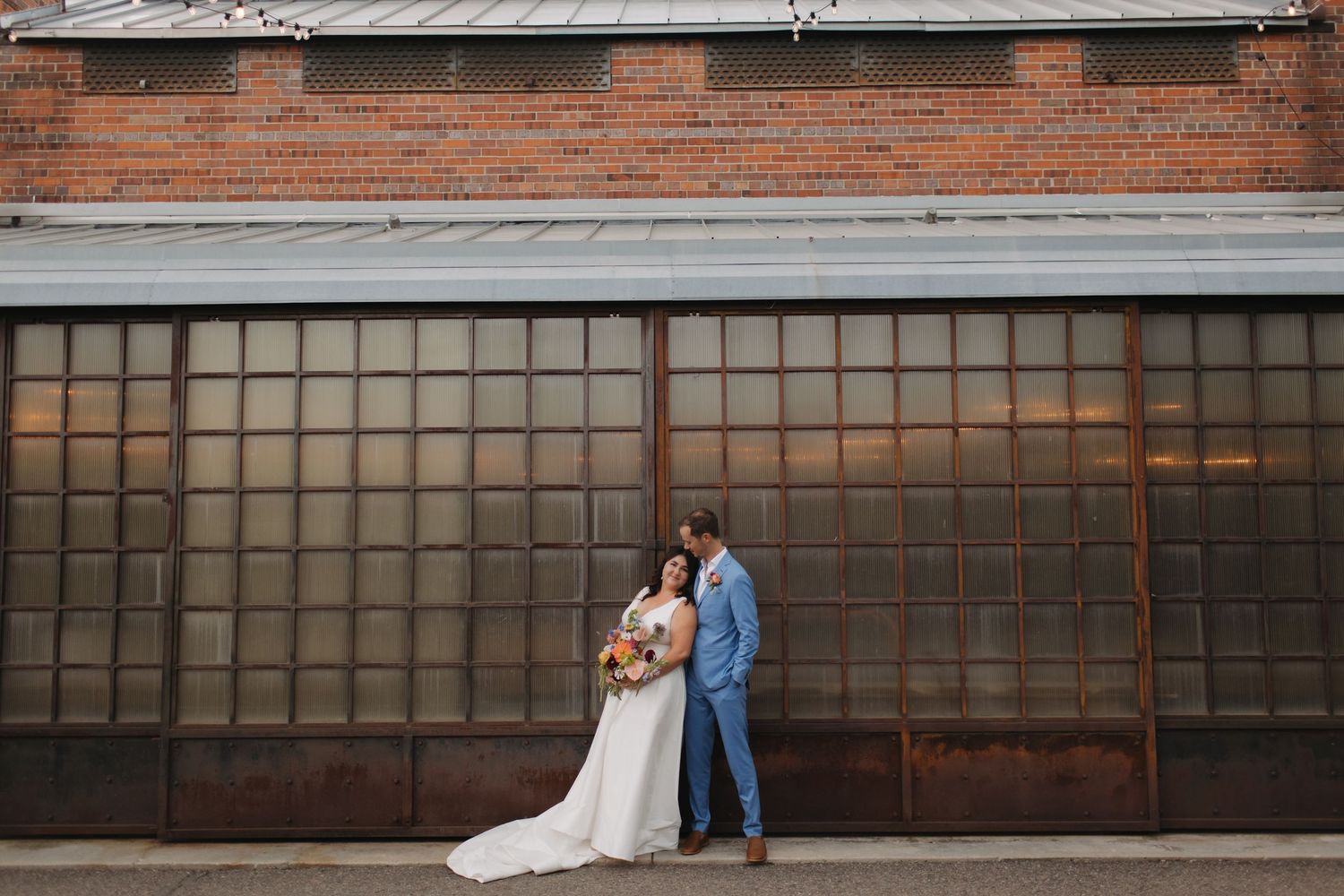Wedding couple poses against industrial brick and window backdrop in urban setting.