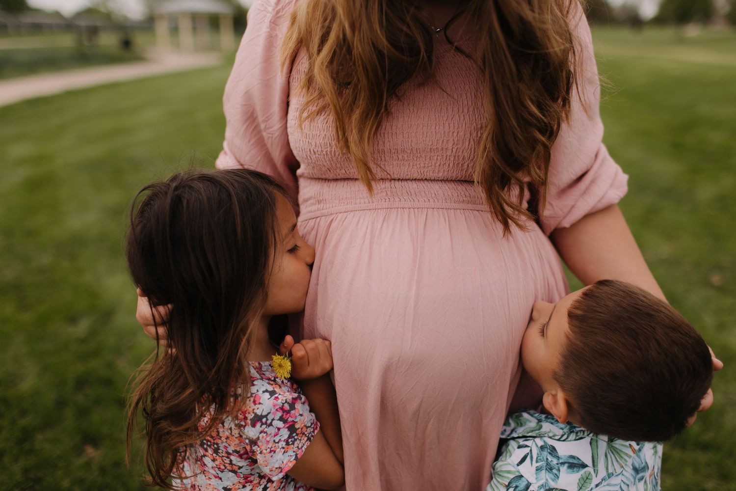 Children hugging pregnant mother wearing pink dress in outdoor setting.