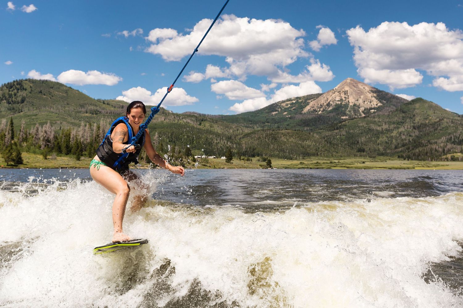 A wakeboarder rides waves on a mountain lake with peaks and blue skies in the background.