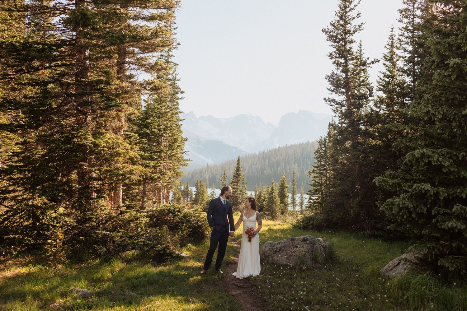 Scenic mountain wedding photography session with evergreen trees and golden sunlight in the background.