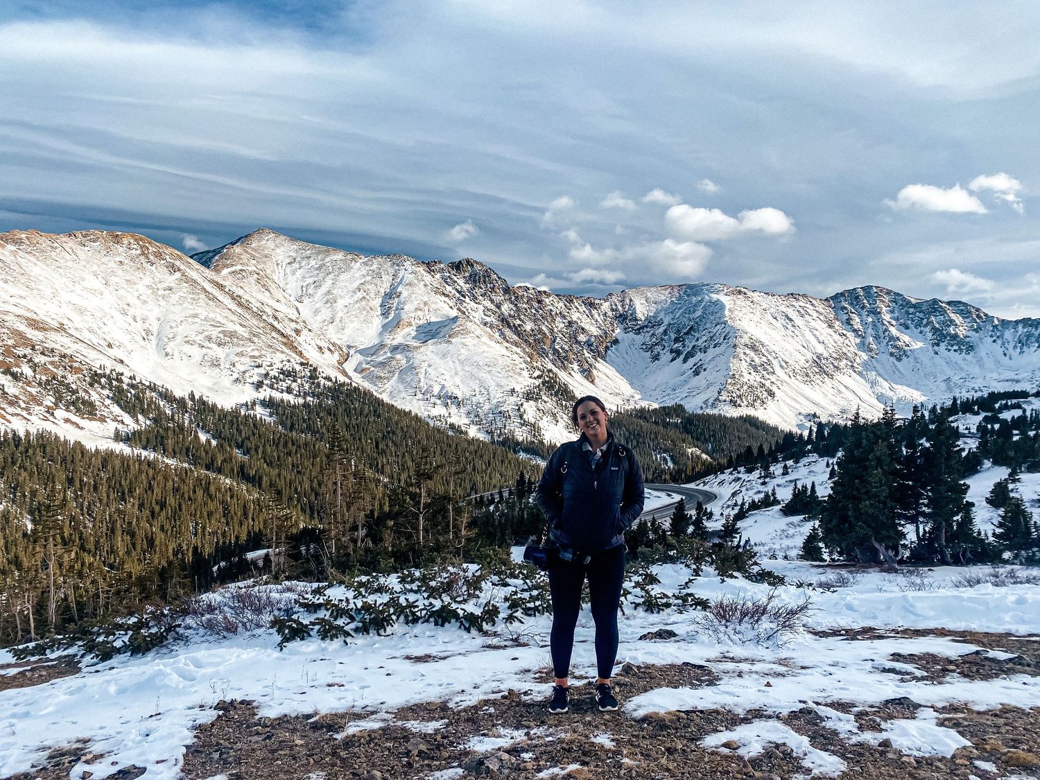 A winter hiker stands on snowy terrain with majestic snow-capped mountain peaks in the background.