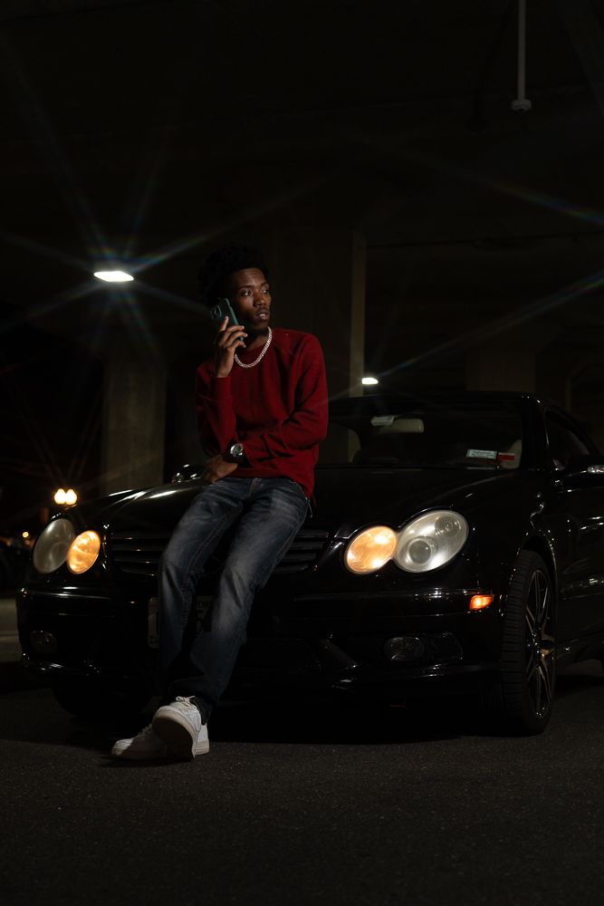 Young man in red sweater and jeans leaning on black Mercedes at night while talking on phone.