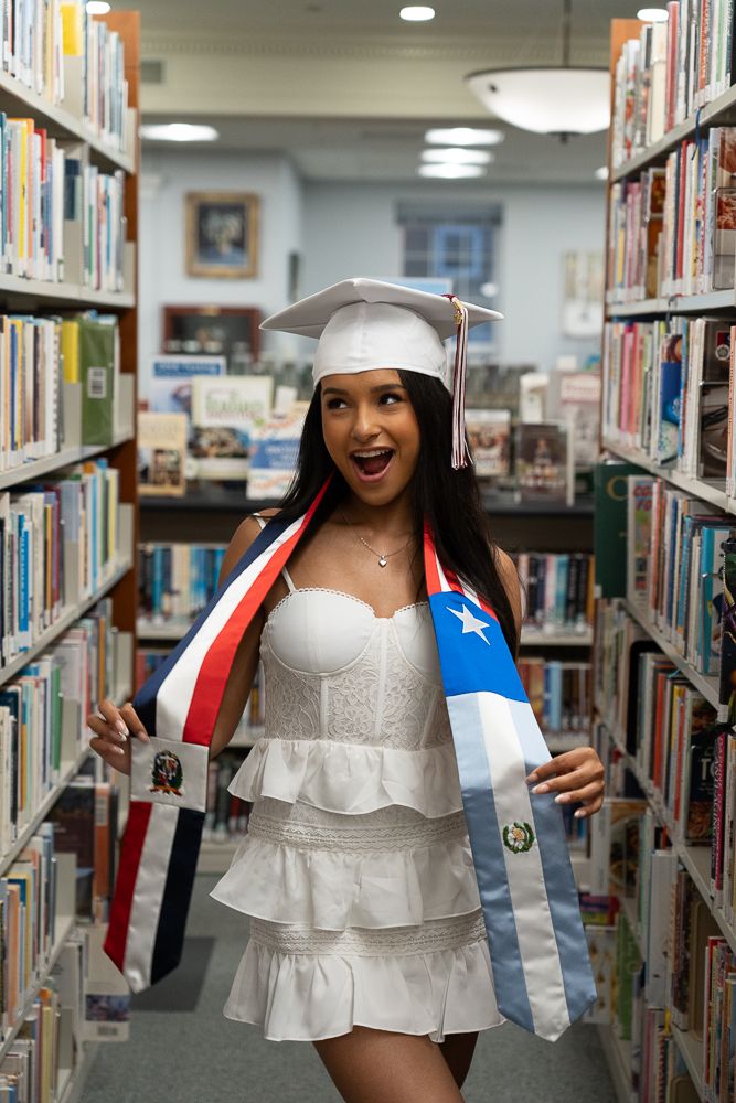 Smiling graduate in white dress and cap poses in library aisle holding Puerto Rican flag sash and diploma.
