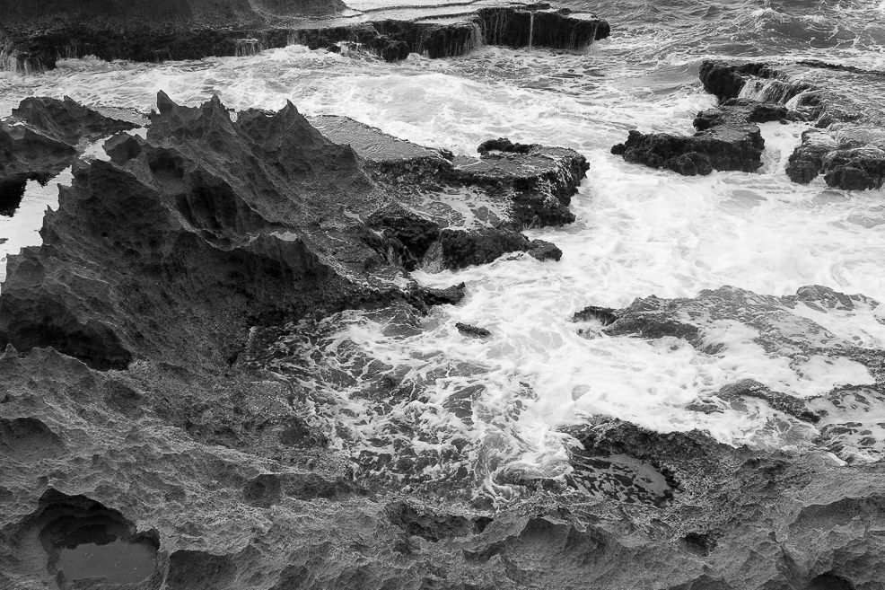 Dramatic black and white seascape showing waves crashing against rocky coastline.