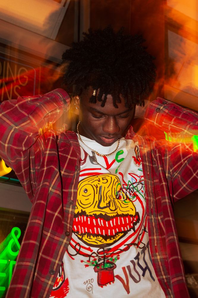 Young man in red plaid shirt and graphic tee poses with hands behind head against colorful neon light background.