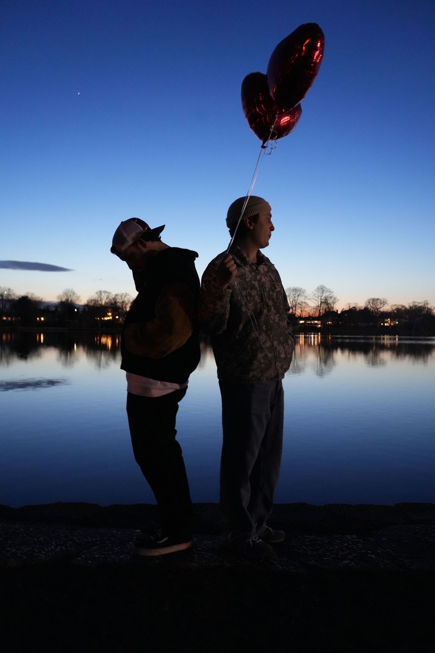 Two figures in silhouette holding balloons against twilight lake view.
