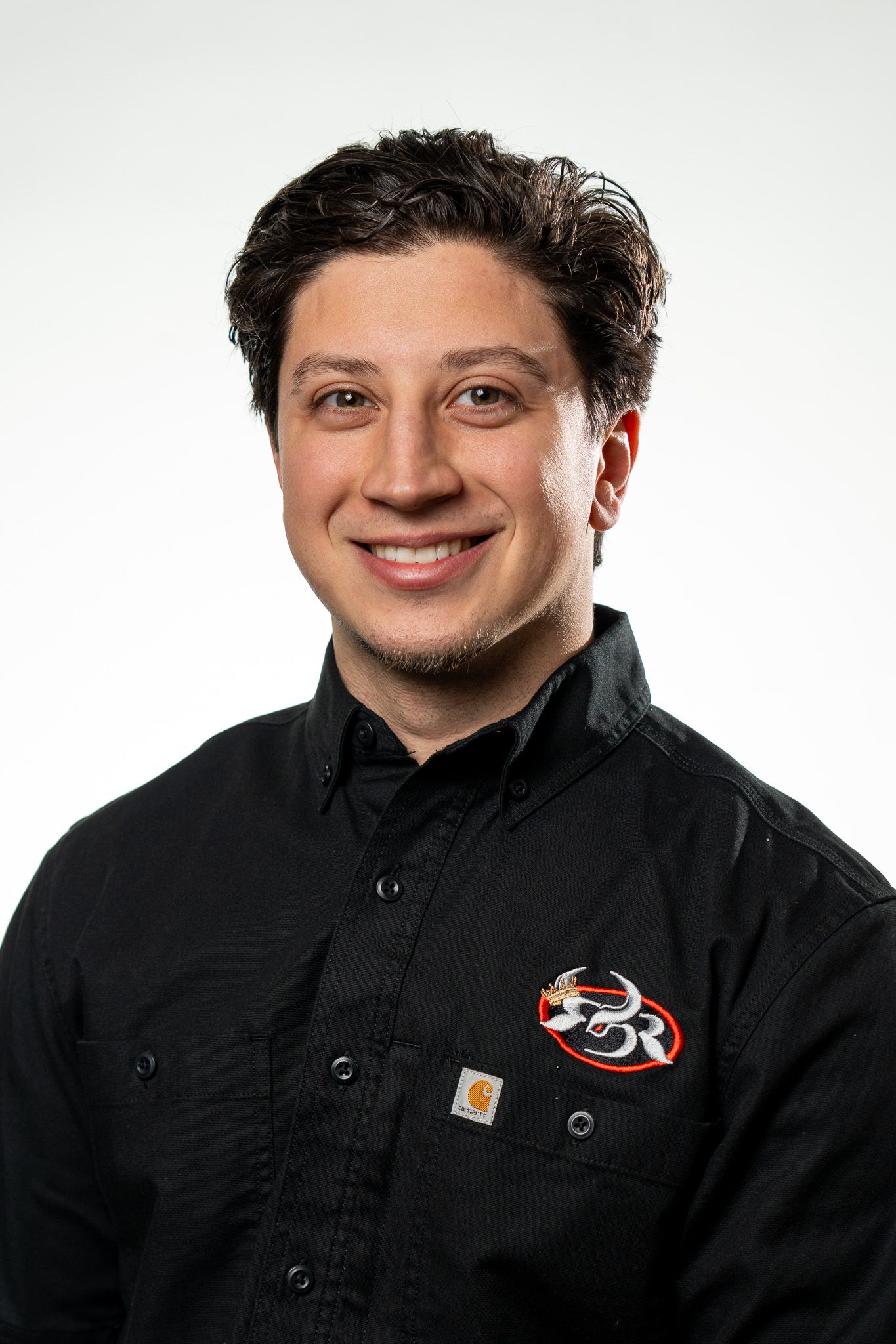 Smiling young man with dark curly hair wearing a black Carhartt button-up shirt with a logo patch on a white background.